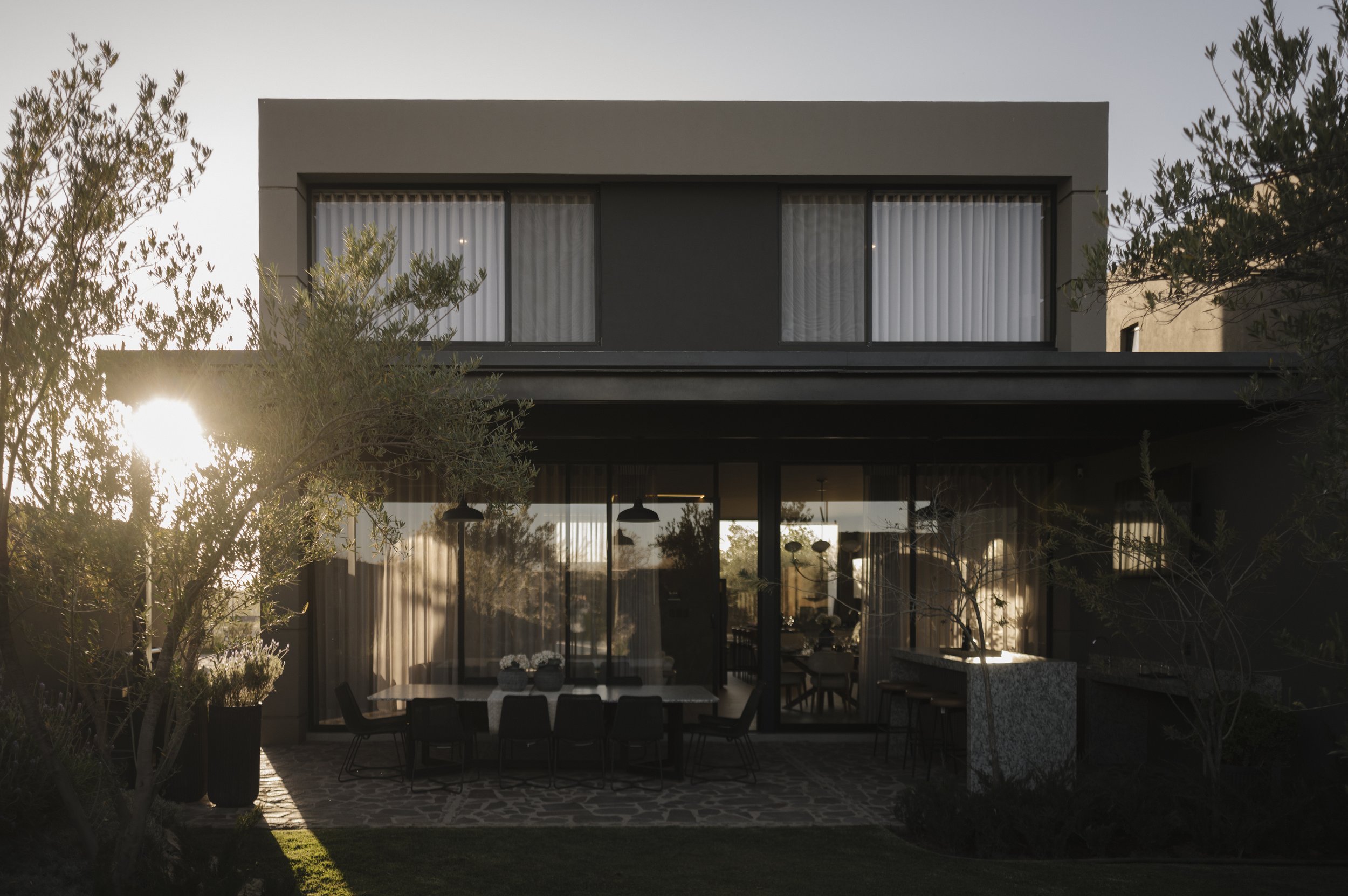 Casa moderna de dos pisos con ventanas grandes, durante el atardecer, con árboles y mobiliario de patio en el primer nivel.