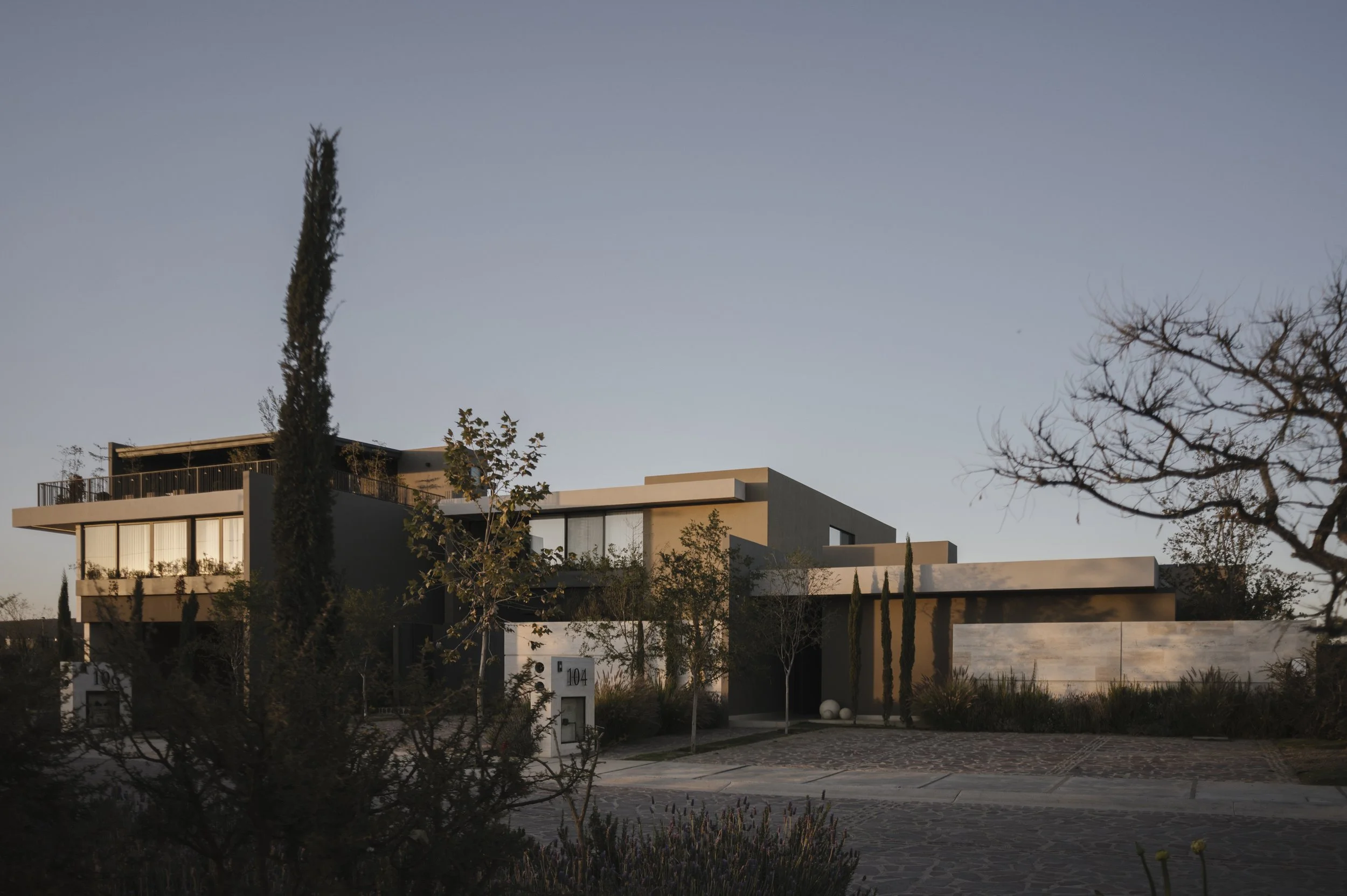 Vista de una casa moderna con varias plantas y ventanas grandes, rodeada de árboles y plantas, durante el atardecer.
