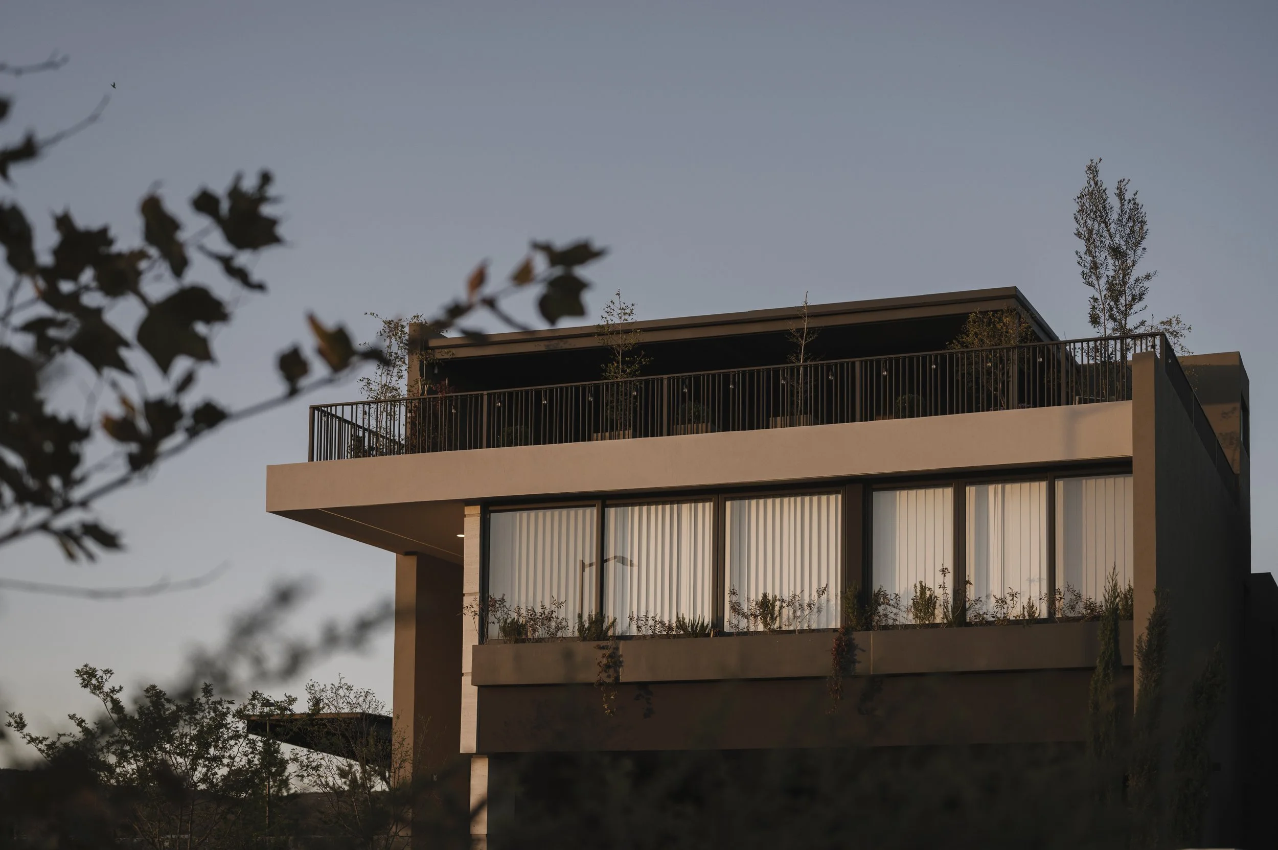 Casa moderna de varios pisos con grandes ventanas y balcón en la parte superior, rodeada de árboles y plantas.