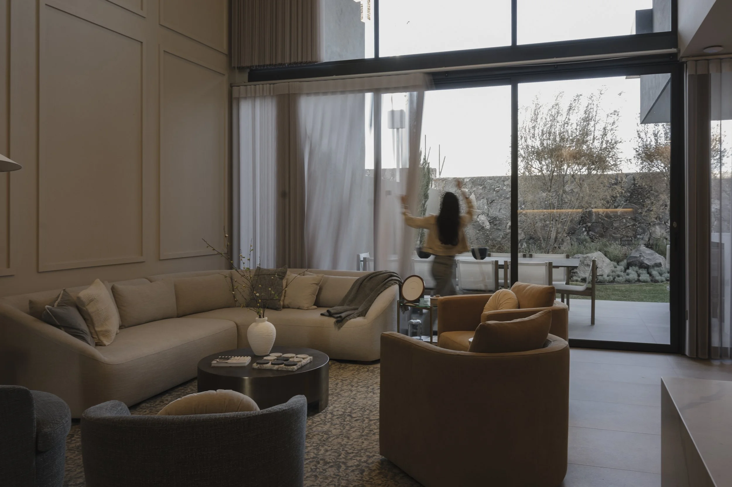 Mujer ajustando la cortina en una sala de estar moderna con sofá beige y sillones, vista hacia un patio exterior con césped y árboles