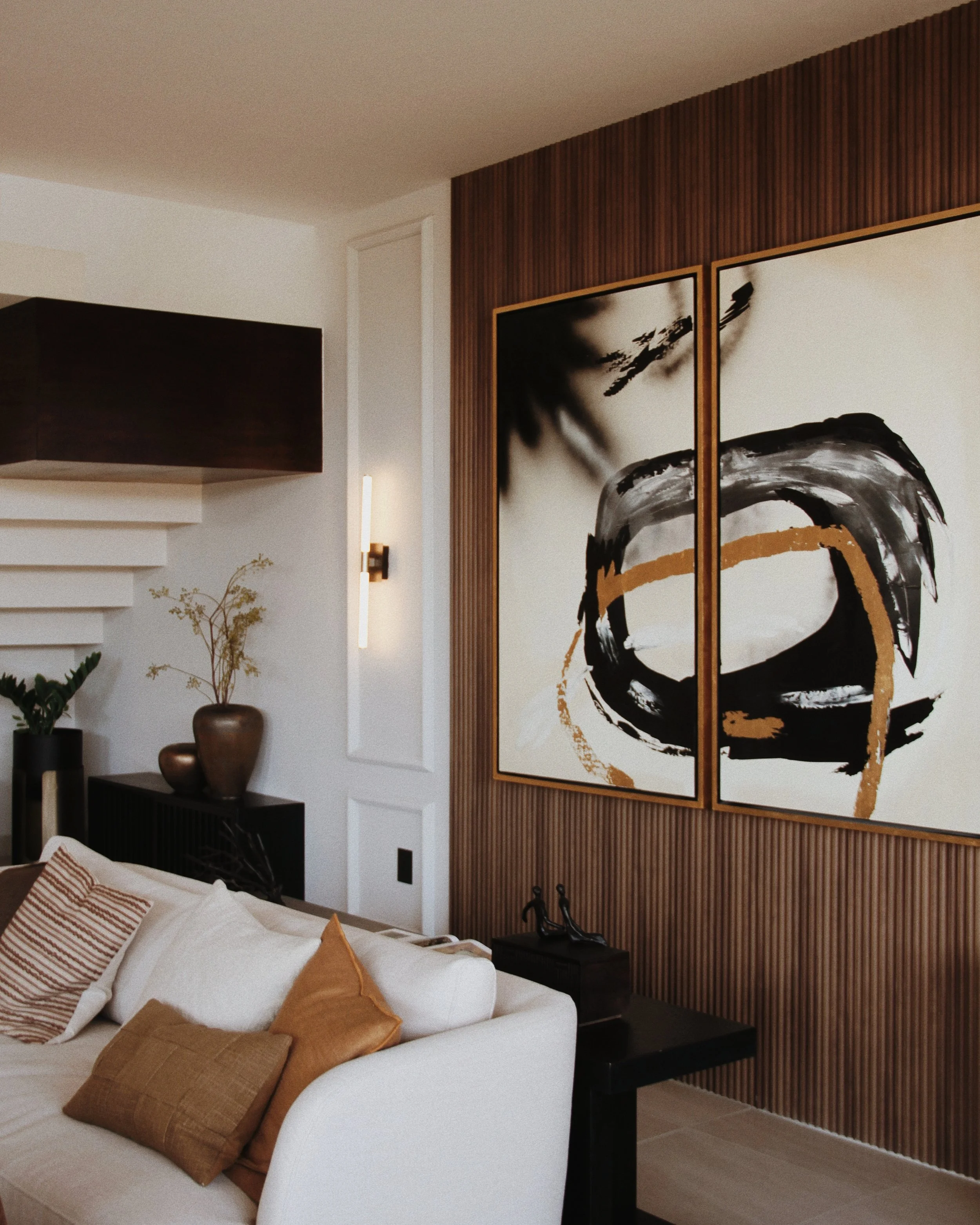 Sala moderna con una pared de madera y arte abstracto en color negro, blanco y dorado, junto a un sofá blanco con cojines en tonos neutros.