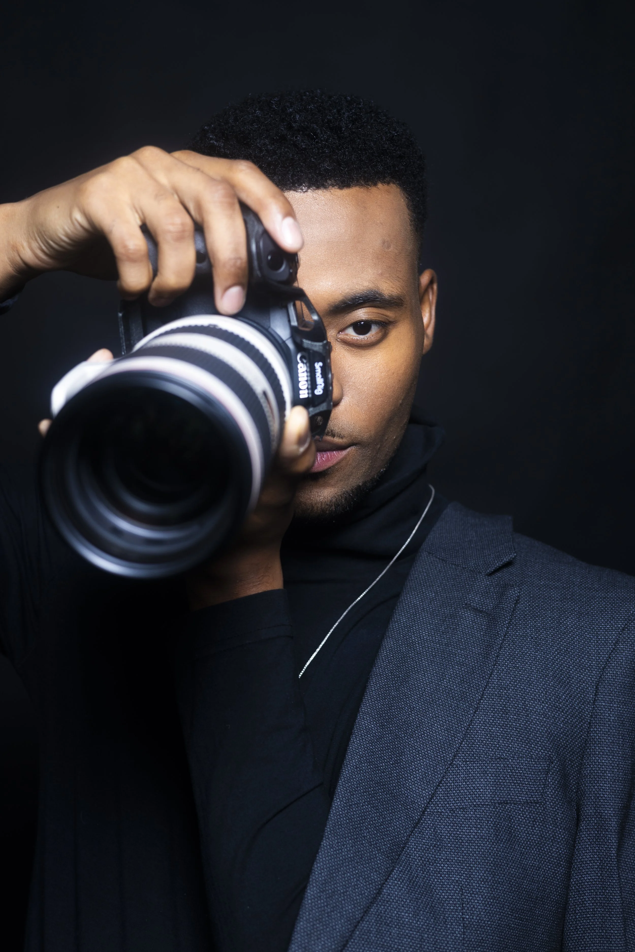Young man with black curly hair holding a camera close to his eye, wearing a dark suit against a black background.