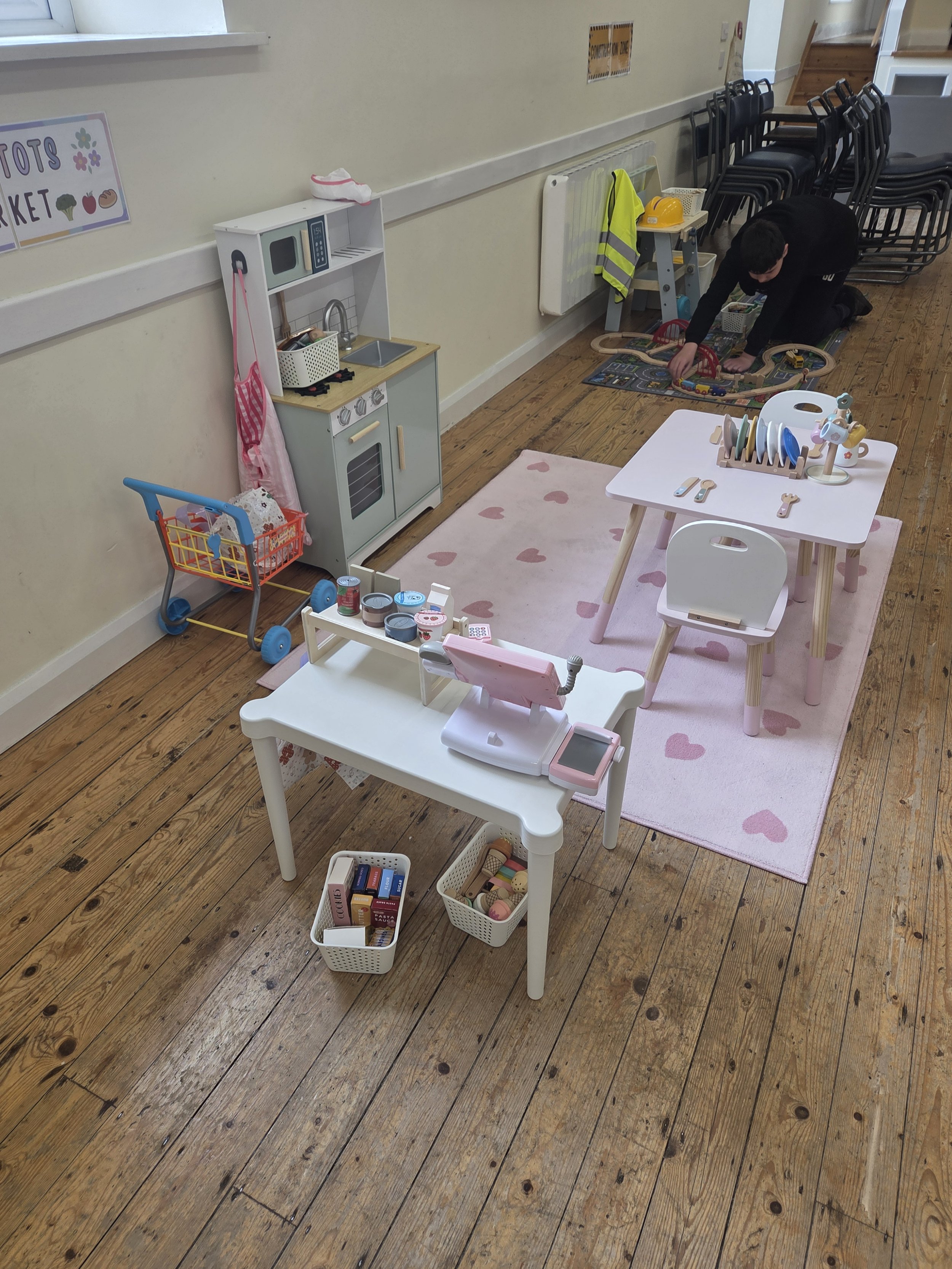 Children's play area with toy kitchen, toy dishes, shopping cart, small table with wooden toys, play food, and a pink rug on wooden floor. A person is playing with a train set on a mat in the background.