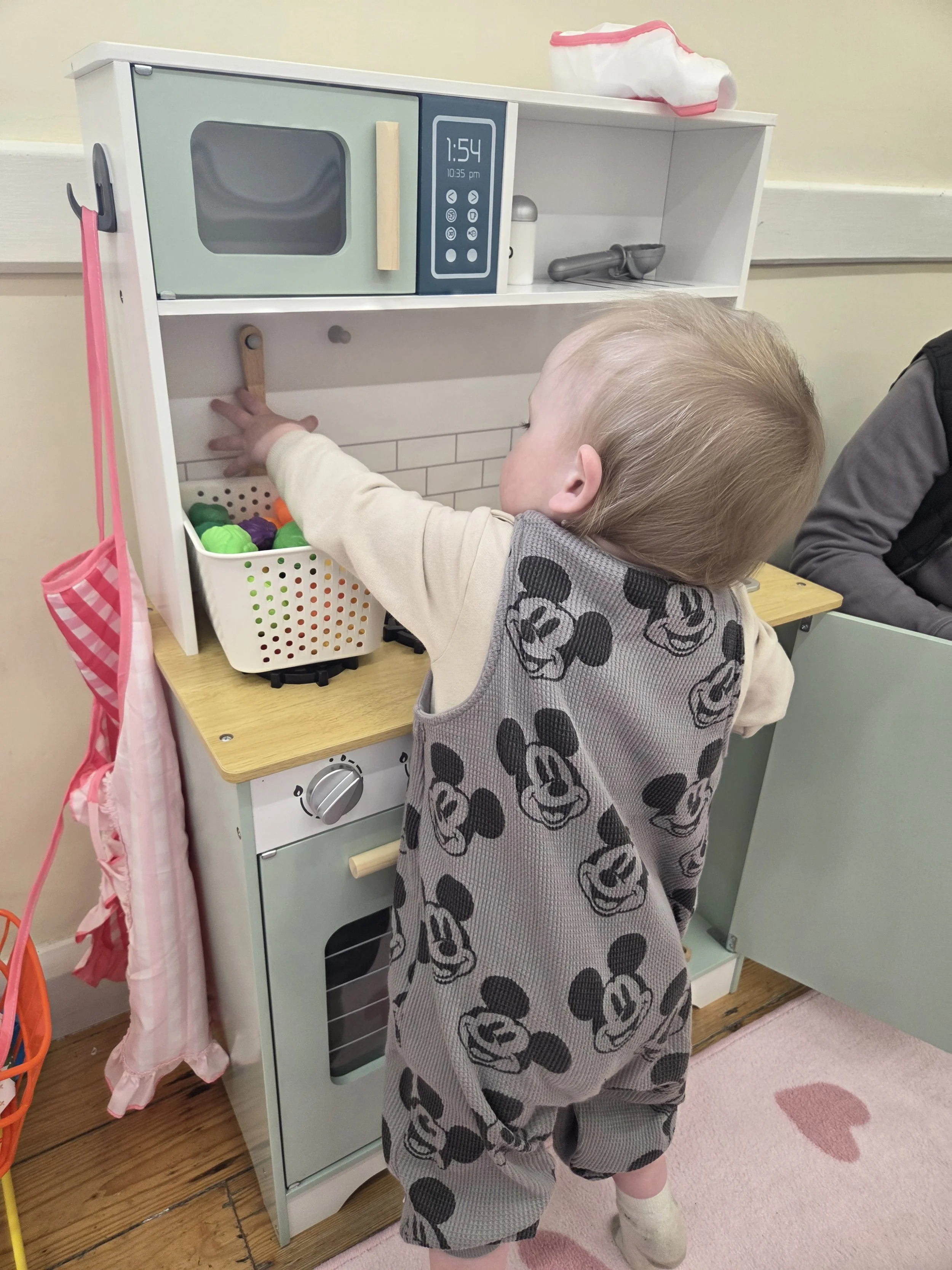 A young child with blonde hair, wearing a Mickey Mouse patterned outfit and beige socks, reaching into a toy kitchen set with a pretend microwave, stove, and utensils. The scene is in a playroom with a pink heart rug and a pink and white apron hanging nearby.