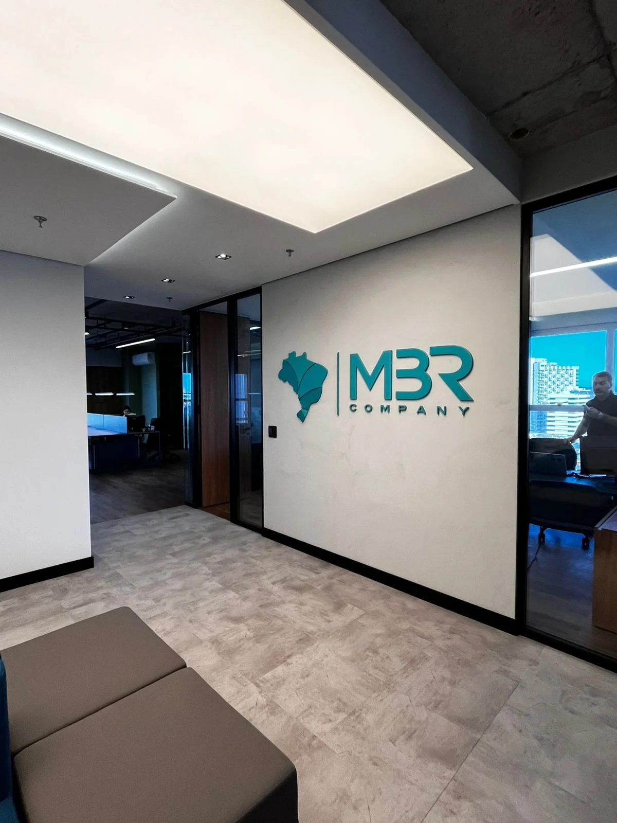 Entrada de escritório com logo da MBR Company na parede, incluindo um mapa do Brasil estilizado, visual moderno com paredes brancas, piso claro e divisórias de vidro.