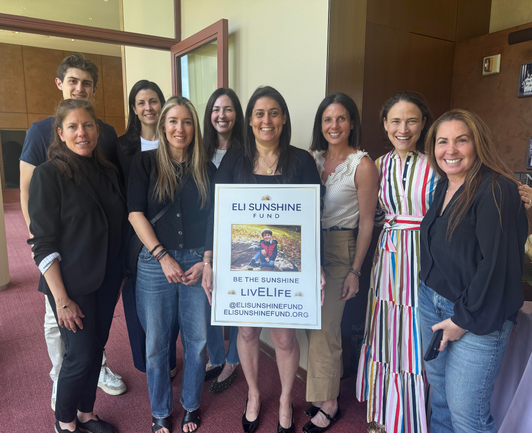 A group of nine smiling people, including seven women and two men, standing indoors holding a sign promoting the Eli Sunshine Fund, which features a picture of a young boy sitting outdoors and messages about being the sunshine and supporting life.
