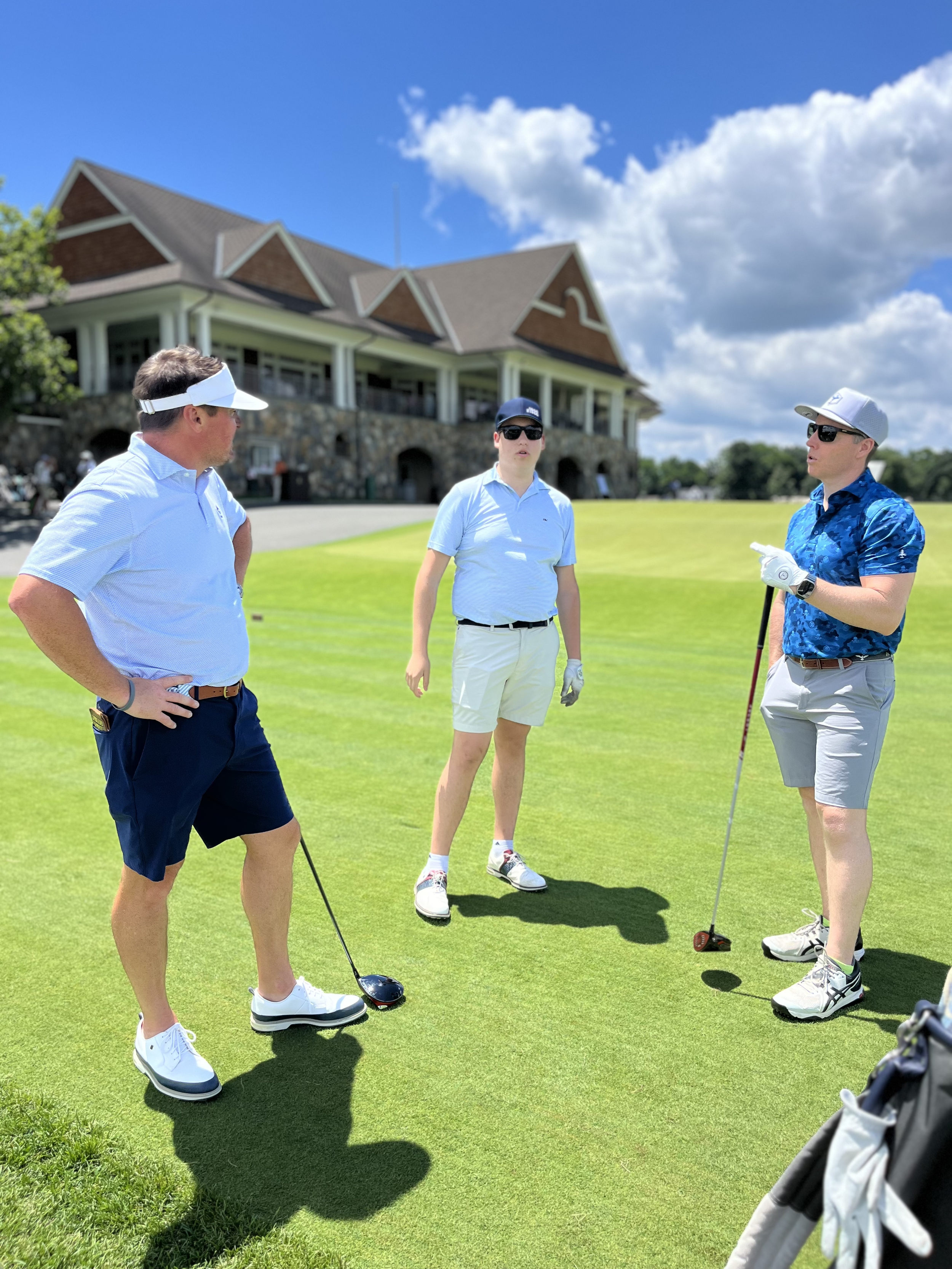 Three men on a golf course standing on the green grass, engaged in conversation, with a building in the background under a partly cloudy sky.