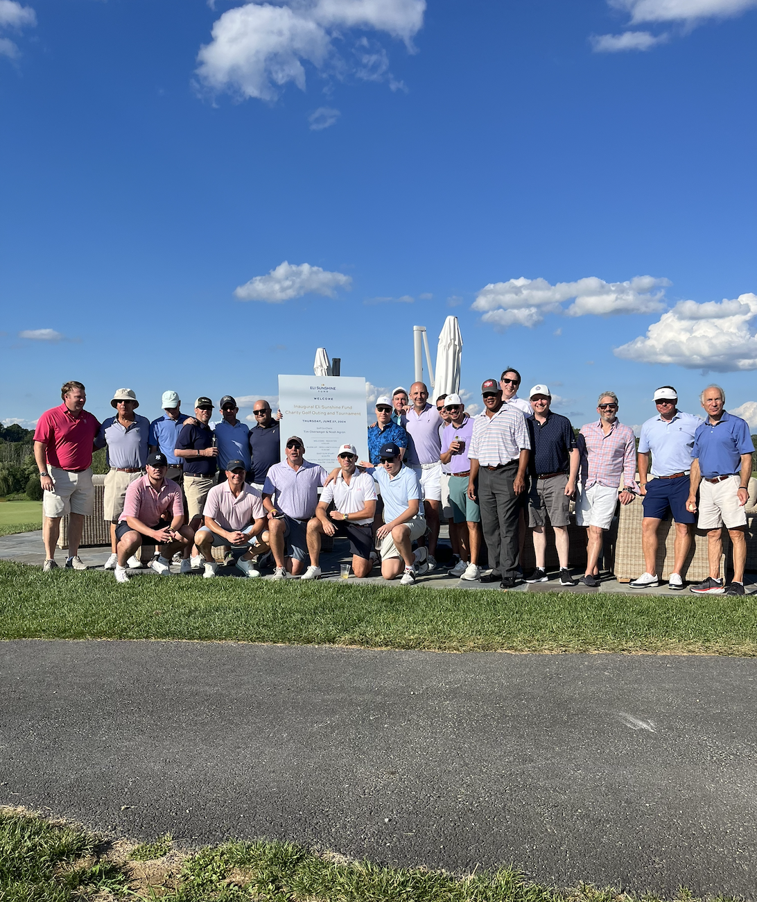 Group of men and women posing outdoors on a golf course during a sunny day, some holding drinks, grouped around a sign and umbrellas.