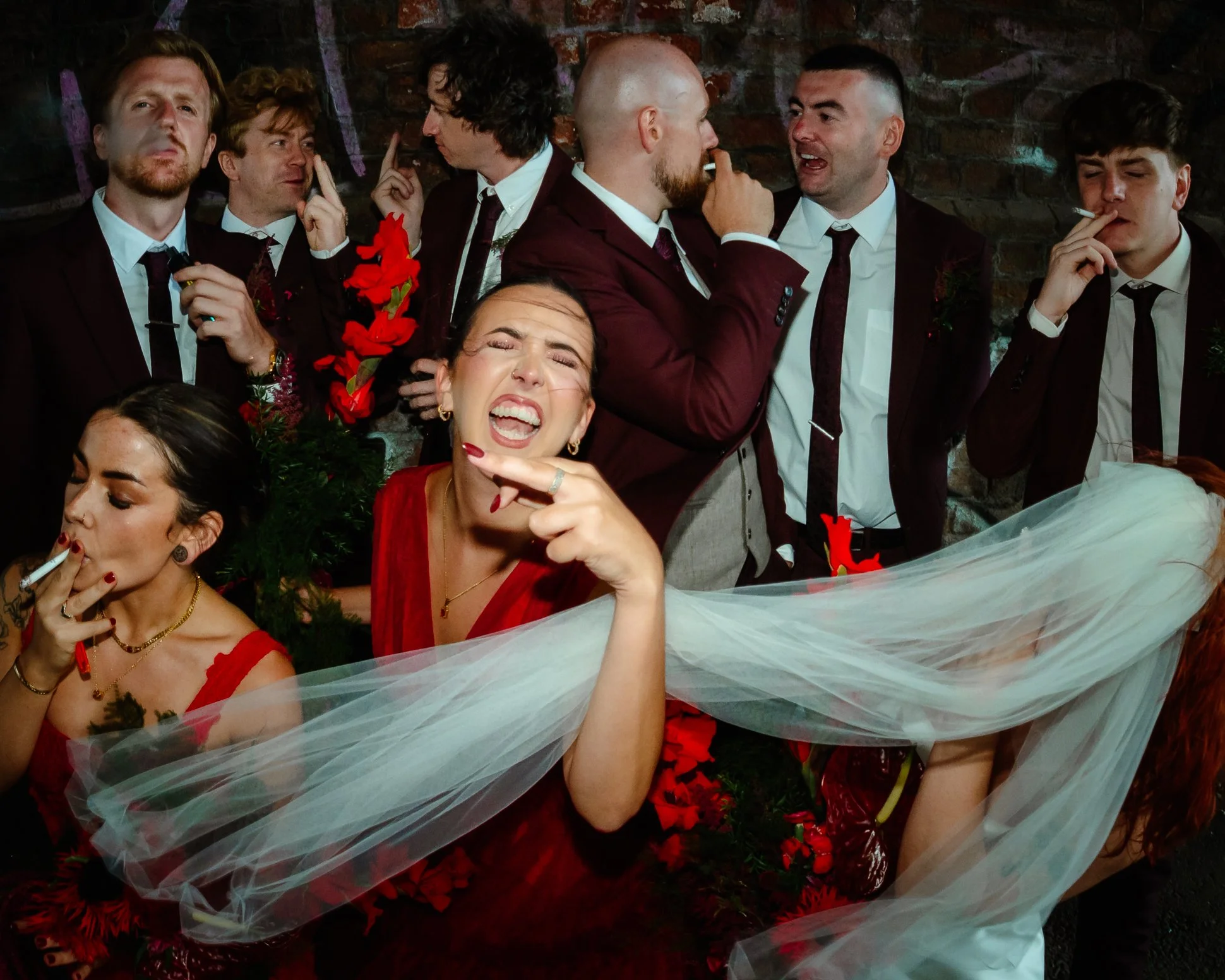 An unserious and wild wedding party. A bridesmaid is swearing at the camera, they're all smoking and the bride's veil is blowing in the wind at Fairfield social club Manchester.