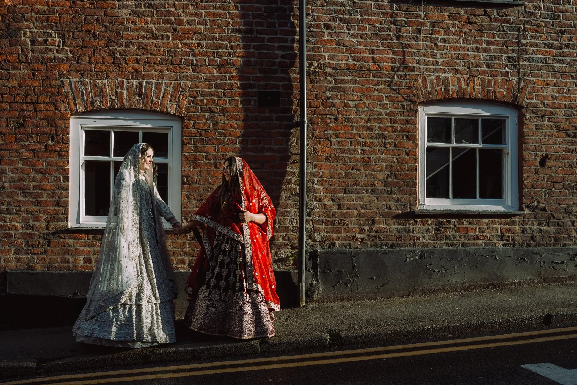 An LGBT couple are walking along a Cheshire street before their wedding. They are a mixed-race couple, and they are both dressed in traditional Asian lehengas. It is a vibrant photo.