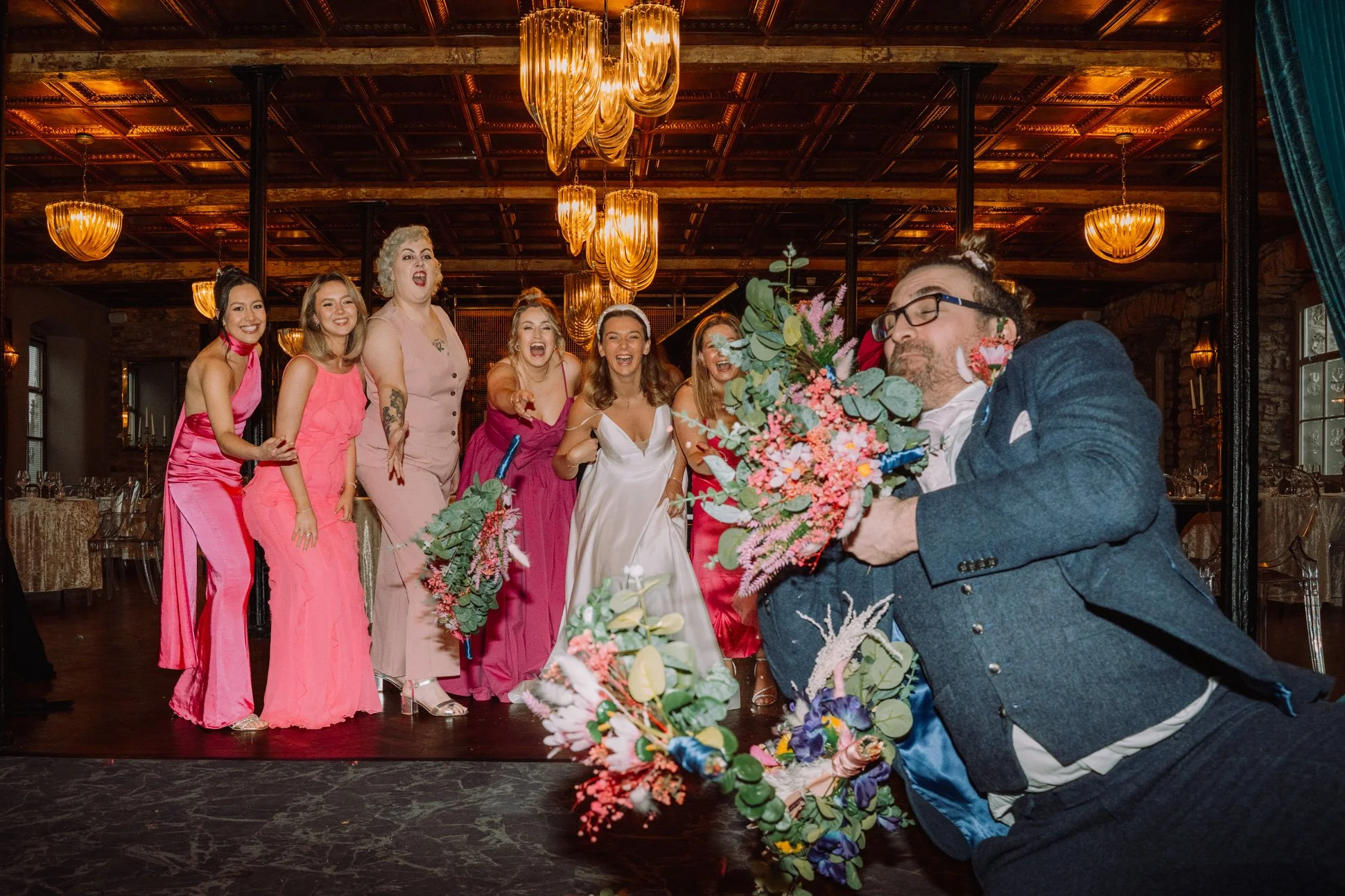 A group of bridesmaids in pink have thrown their bouquets and a groomsman has leapt in to catch them,.