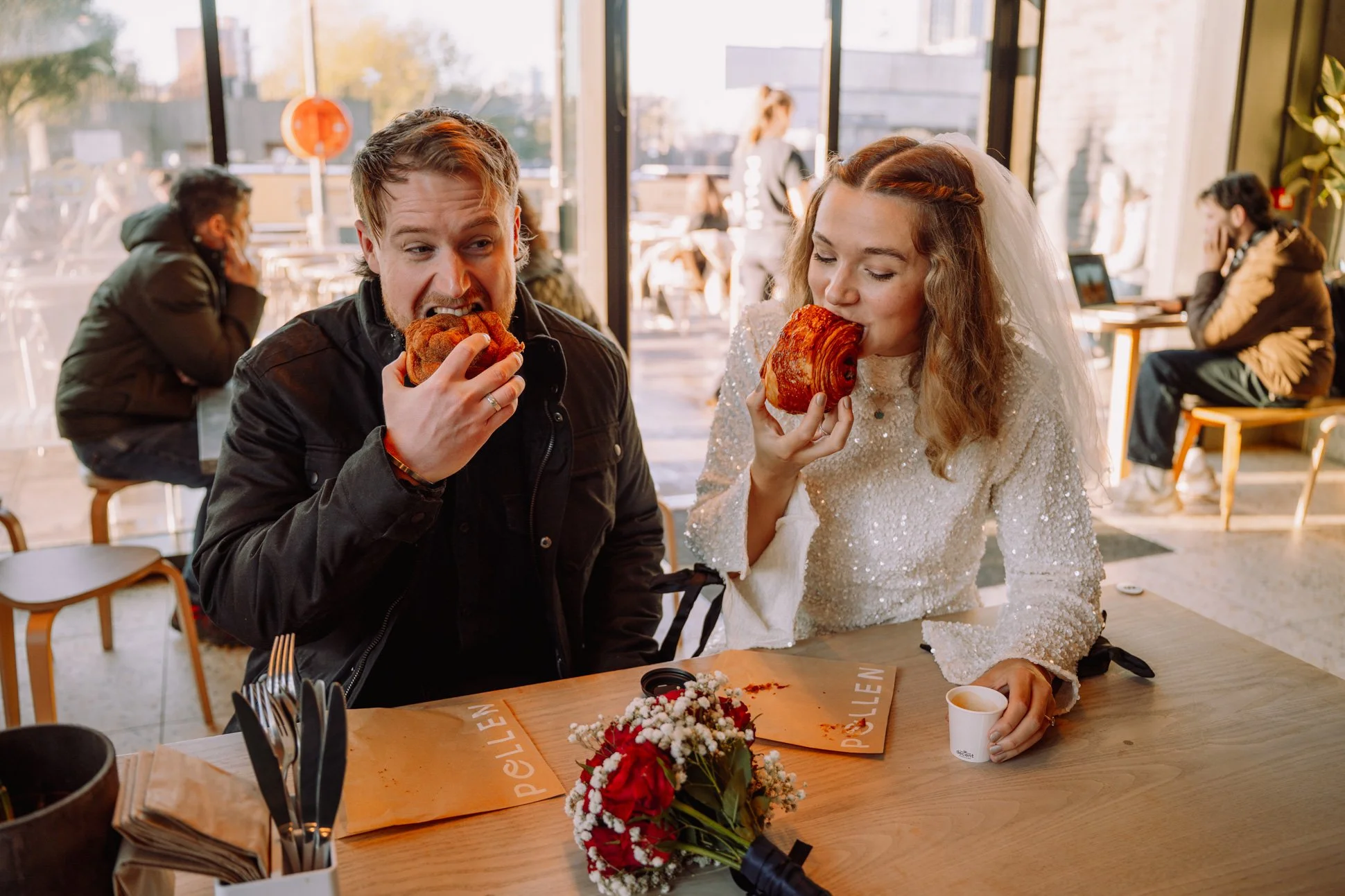 A couple on their wedding day are eating pastries in Manchester, the bride is wearing a mini dress and veil. There are red roses on the table.
