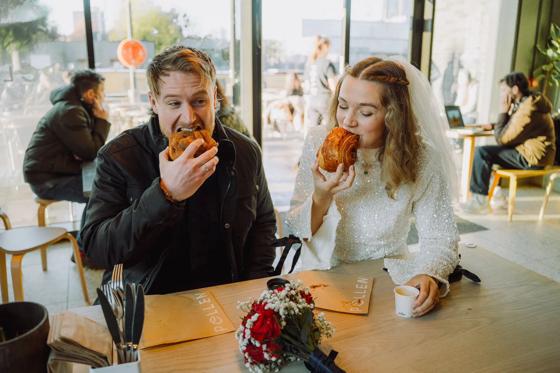 A couple in cool wedding clothes are eating pastries in Manchester before their city centre elopement.