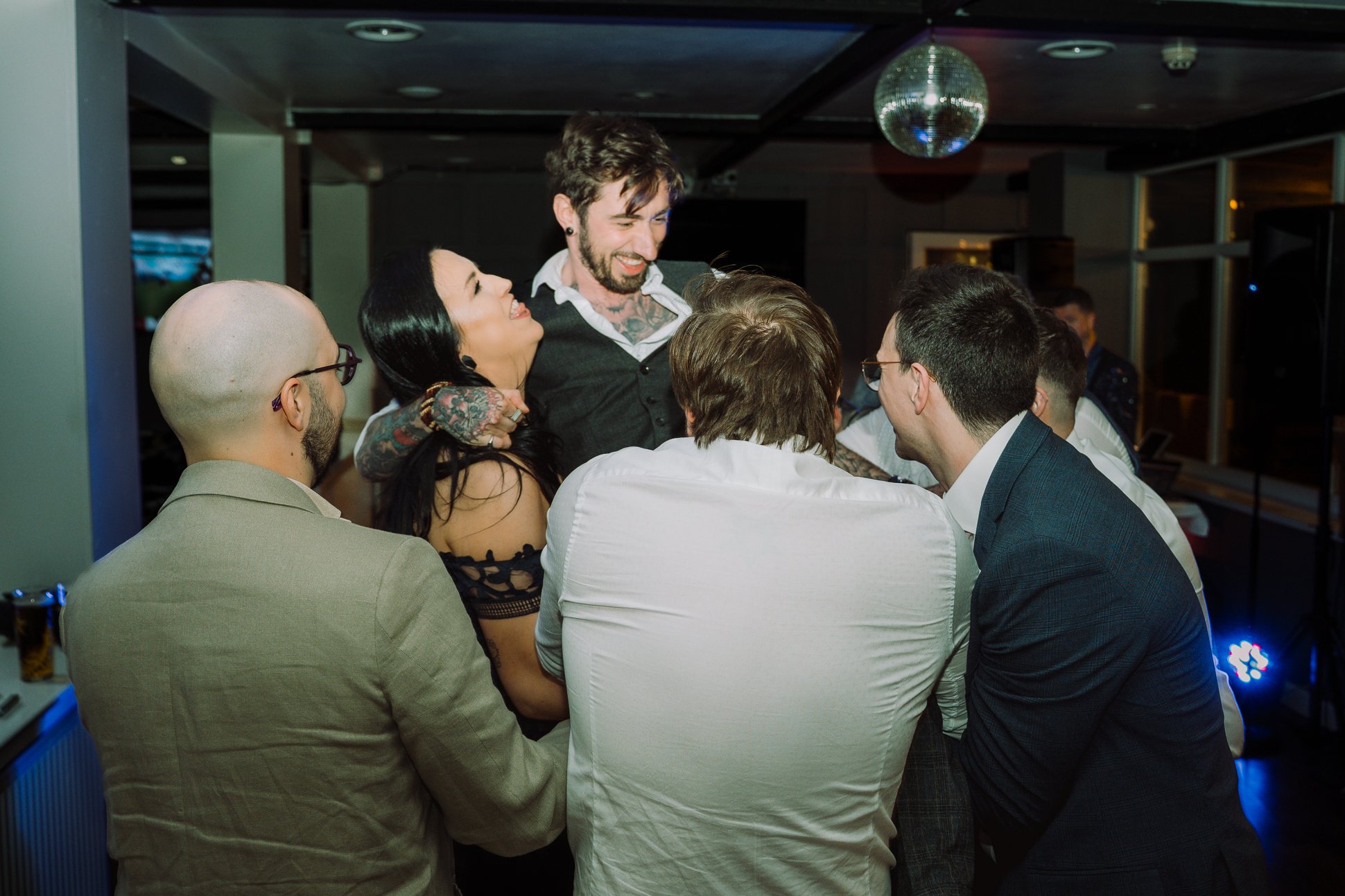 The groomsmen and a tattooed female wedding guest are lifting a tattooed groomsman