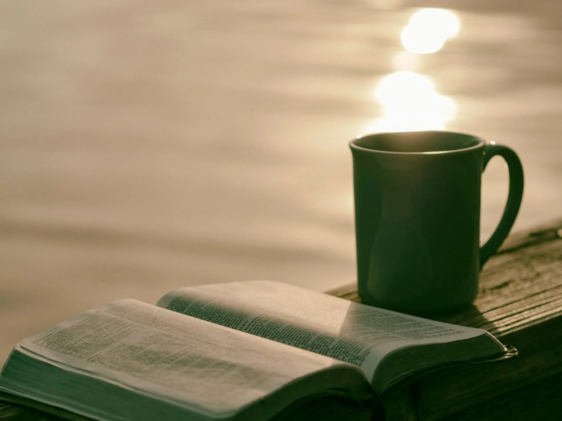 An open Bible, a green mug, and a body of water reflecting sunlight, placed outdoors on a wooden surface.
