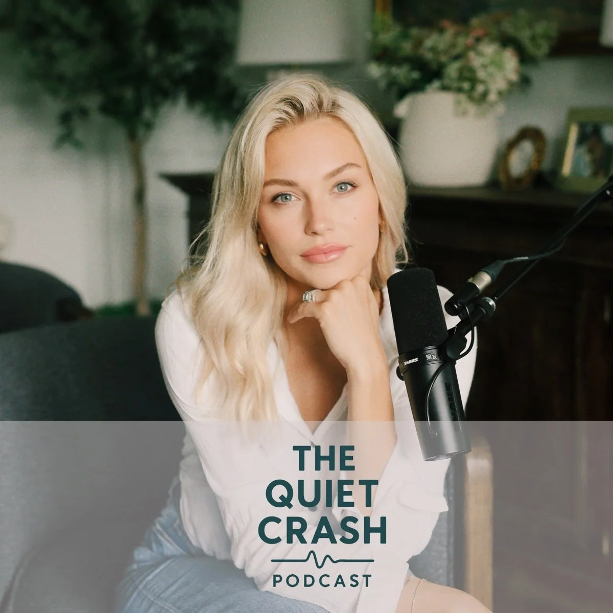 A woman with long blonde hair and blue eyes sitting in front of a microphone in a cozy room with plants in the background. She is resting her chin on her hand and looking at the camera. The text overlay says 'The Quiet Crash Podcast.'