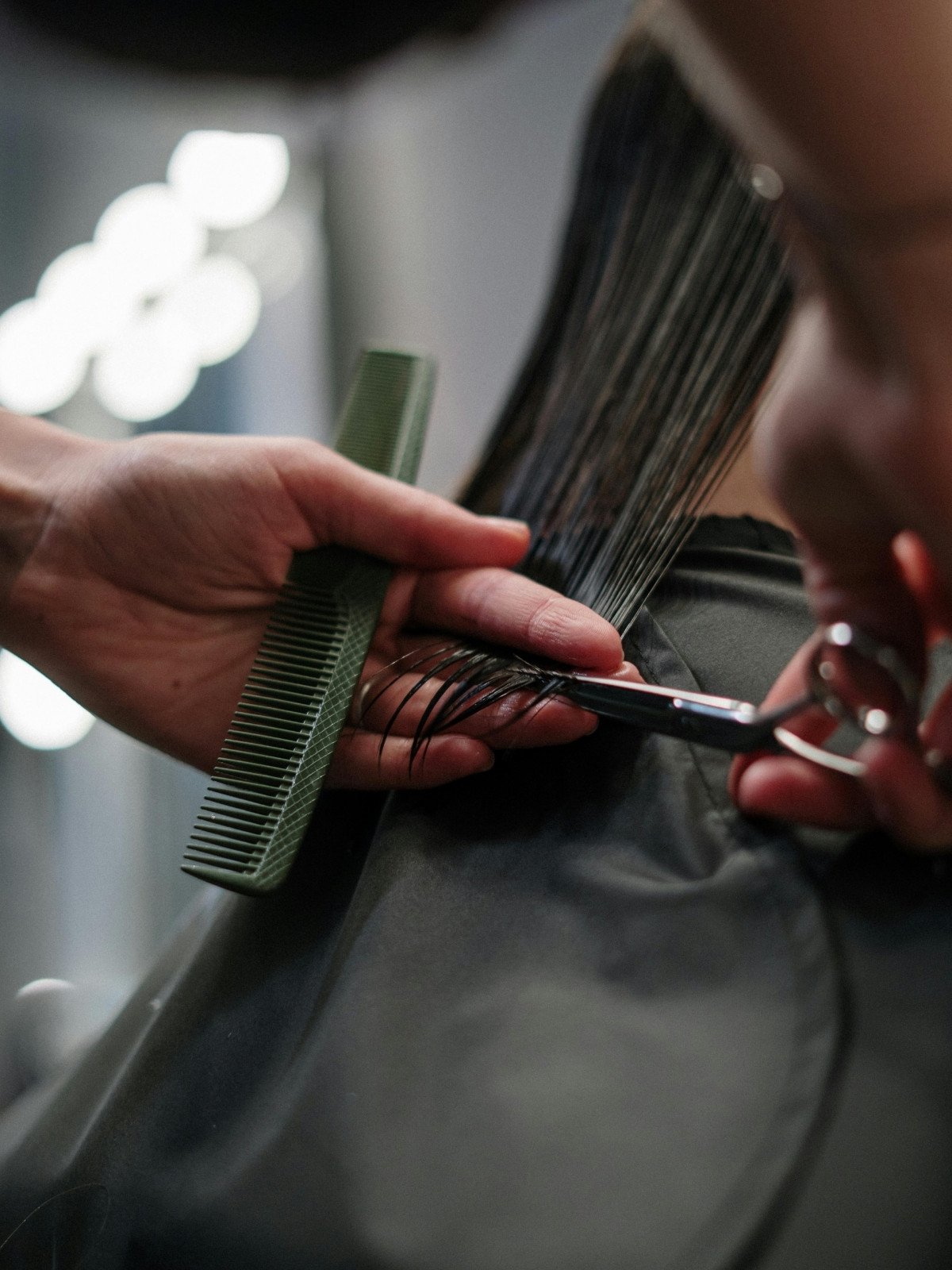 A hairstylist trims a client's wet long hair with scissors, holding the hair with a black sectioning clip and comb in a salon.