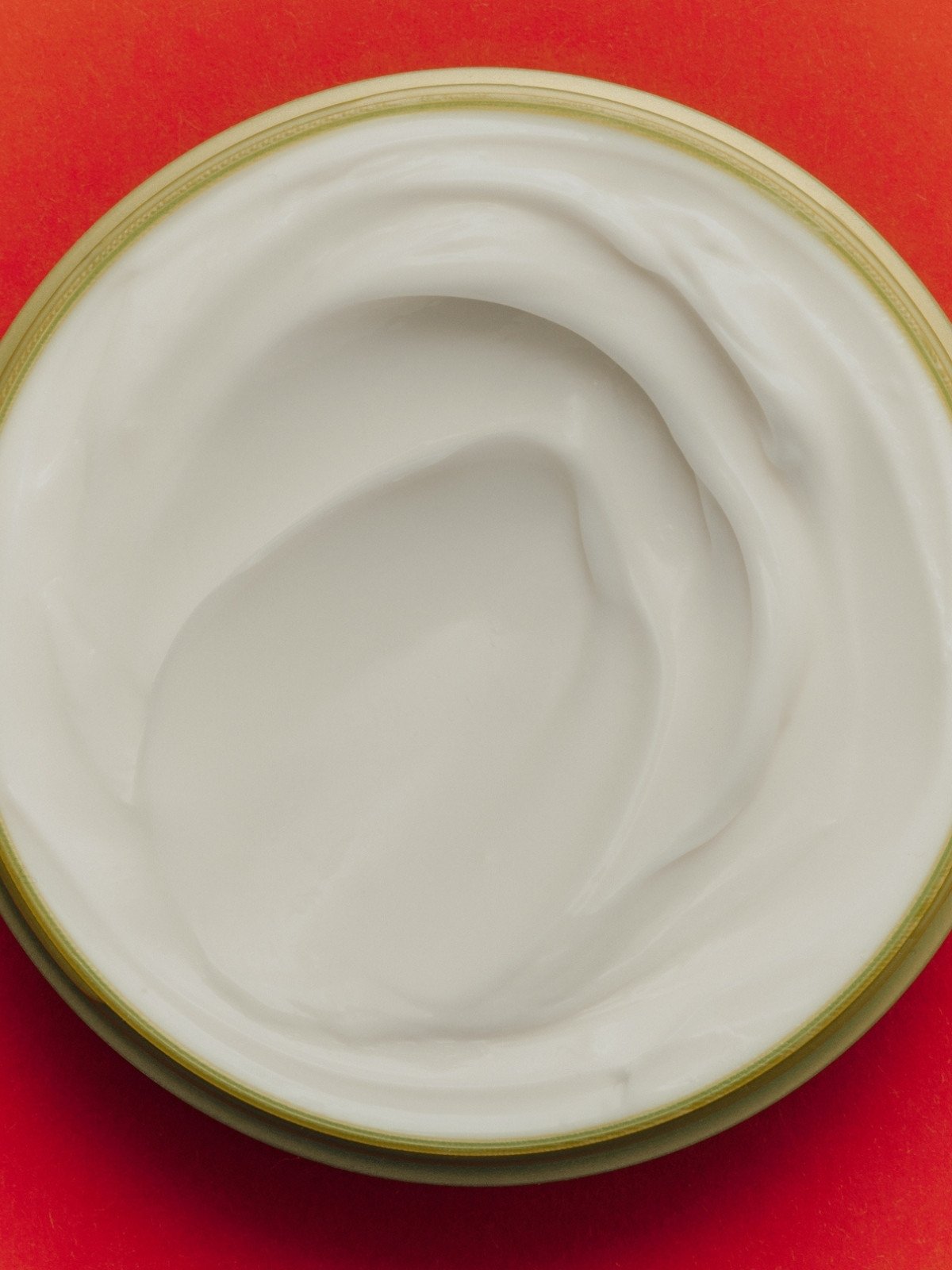White cream or yogurt inside a metal container with a gold rim, placed on a red background.