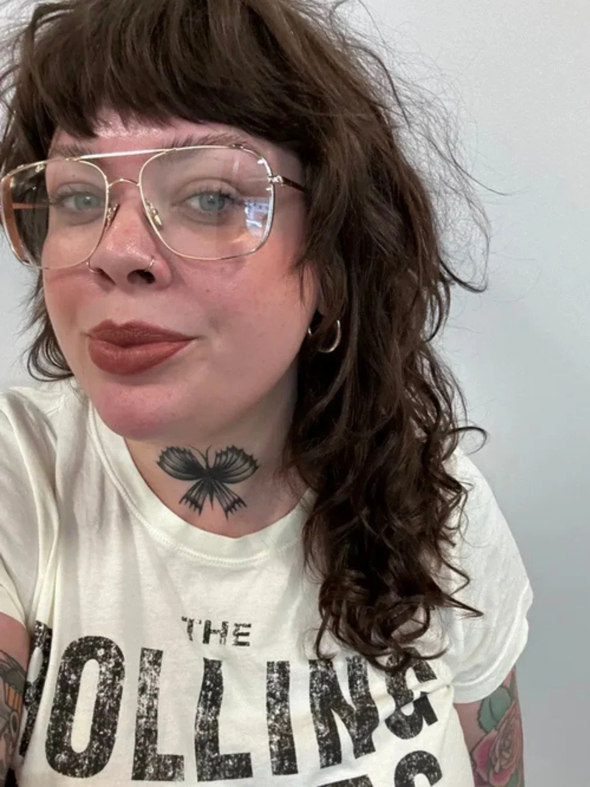 A woman with wavy brown hair wearing large glasses with clear lenses, a white T-shirt with black text, a nose ring, earrings, and tattoos, including a black butterfly on her neck.