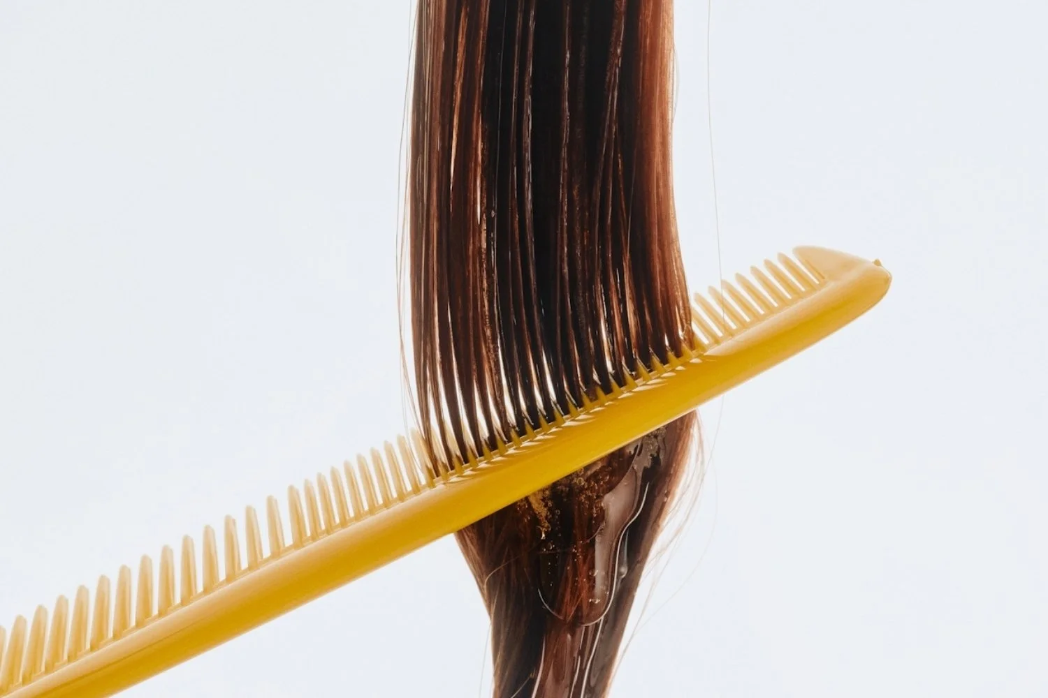 A yellow hairbrush with a comb-like design gently holding a section of wet, brown hair, with hair product or water applying a shiny, slick appearance.