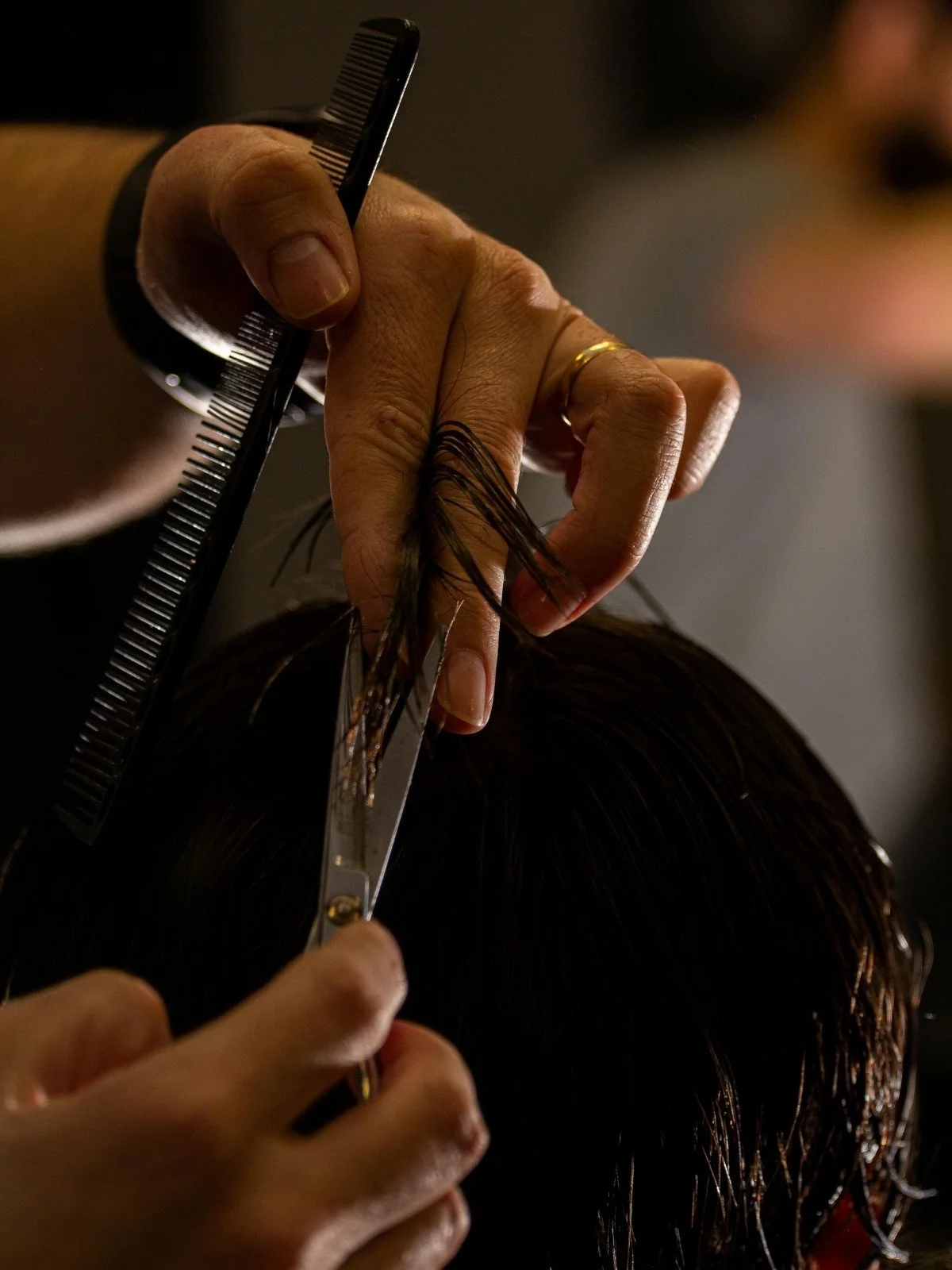 A hairstylist is cutting a client's dark hair with scissors, holding a section of hair between their fingers and using a comb for precision.