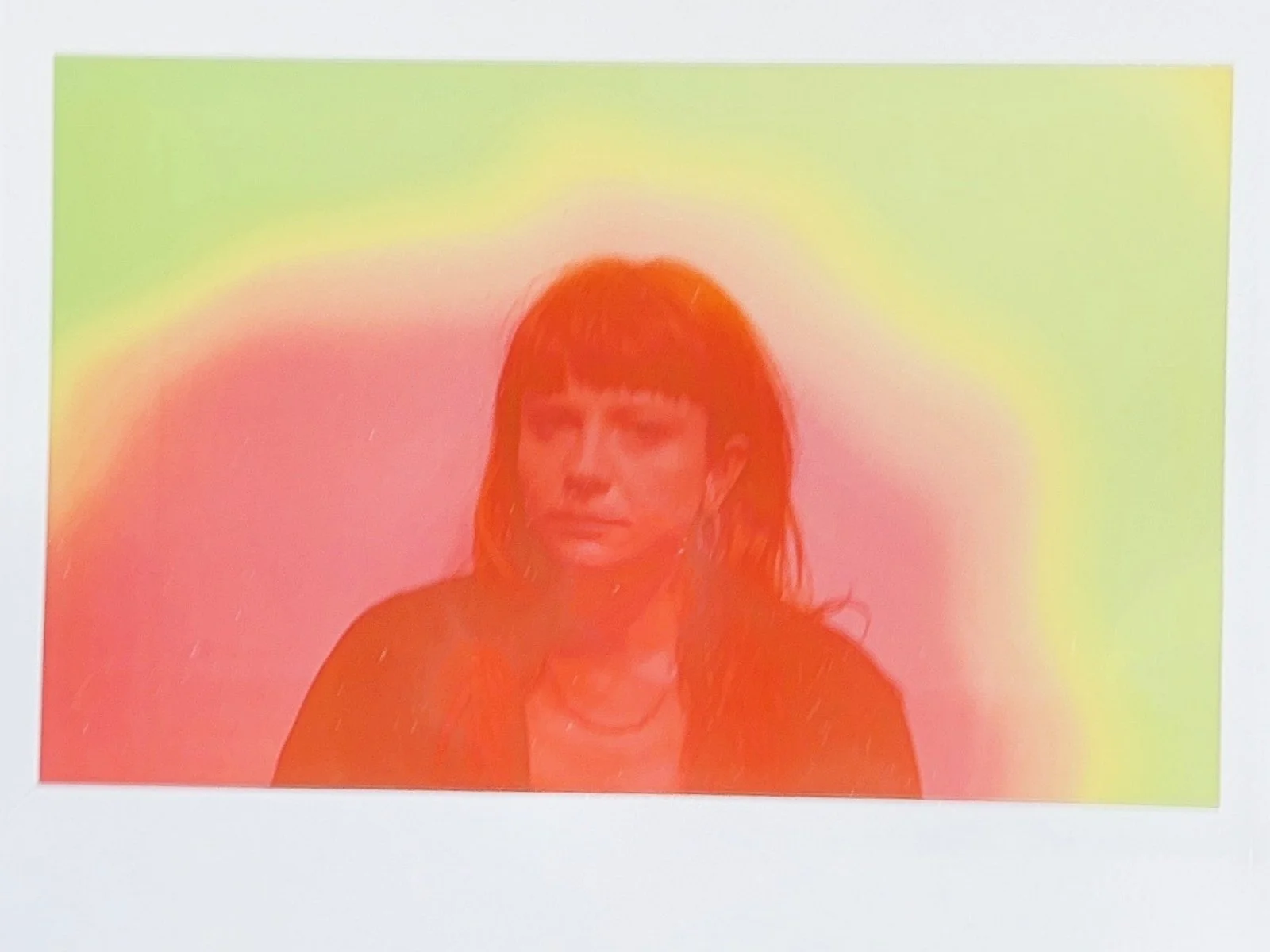 A woman with dark hair and bangs wearing earrings and a necklace, against a colorful background with a gradient of yellow, green, and pink hues.