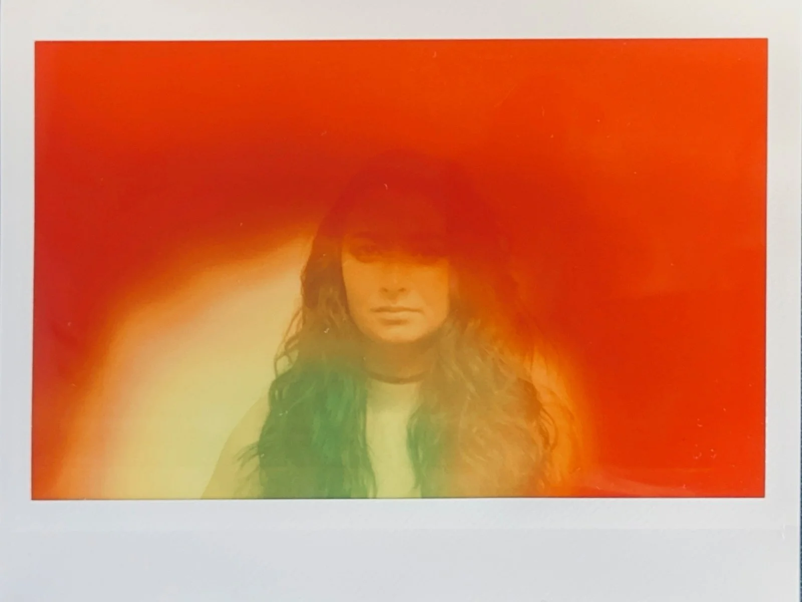 A person with long hair, wearing a white shirt and a dark jacket, standing against a gradient orange background.