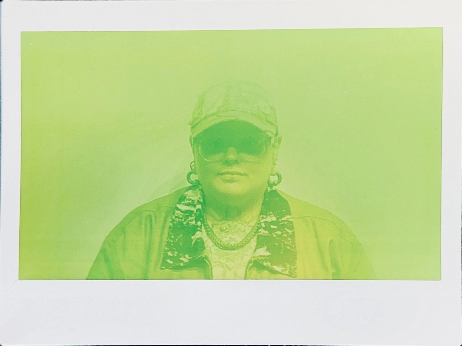 Polaroid photo of a woman wearing sunglasses, a hat, earrings, and layered clothing, displayed on a neon green background.