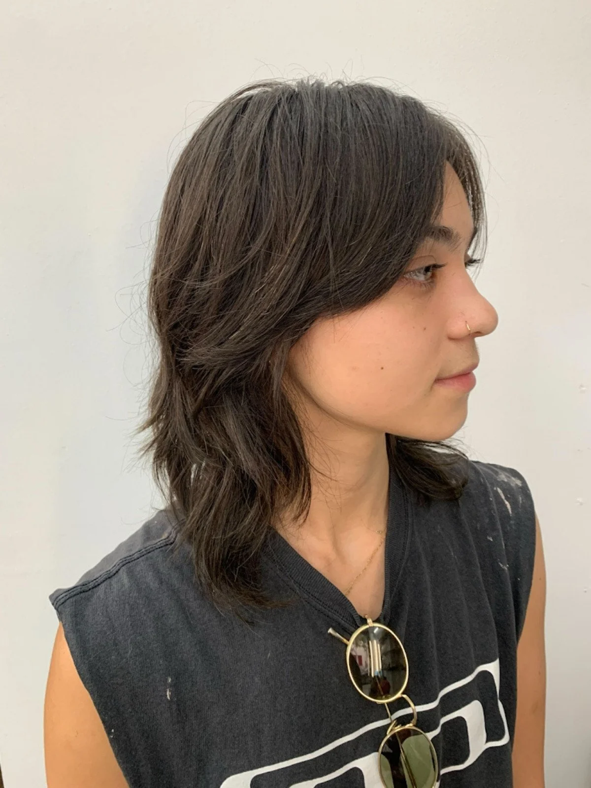 A woman with shoulder-length wavy dark brown hair, wearing a sleeveless black top and sunglasses hanging from a necklace, looking to her right against a plain white background.