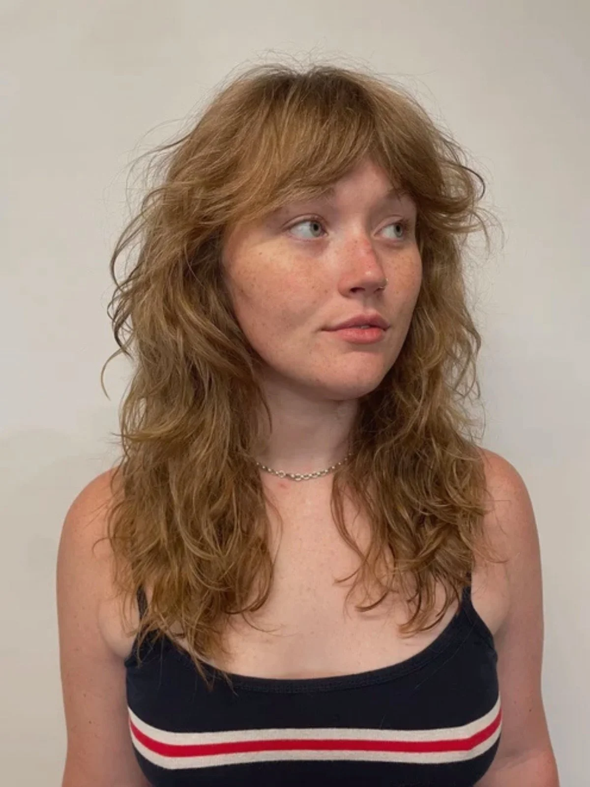 A young woman with curly red hair, light skin, and freckles, wearing a black sleeveless top with red and white stripes, a silver necklace, and a nose ring, standing against a plain white background.