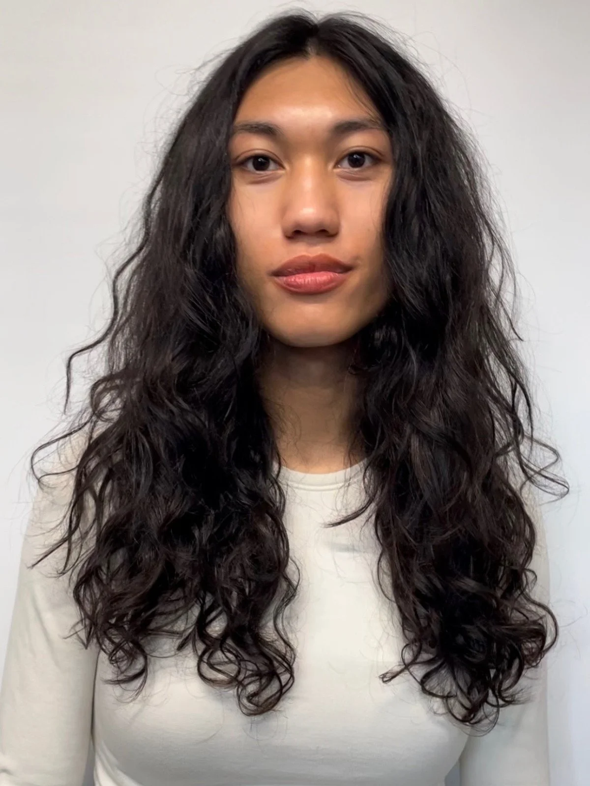 A woman with long, dark, curly hair and wearing a light-colored top, standing against a plain background.