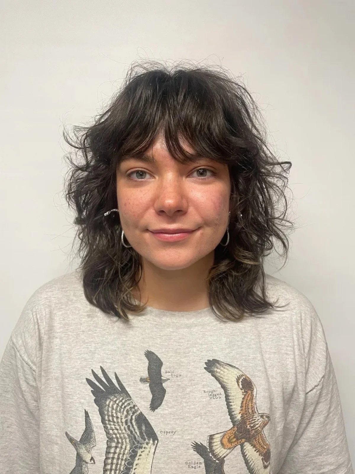 A woman with shoulder-length curly dark hair, wearing a light-colored sweatshirt with images of different bird species, standing against a plain off-white wall.