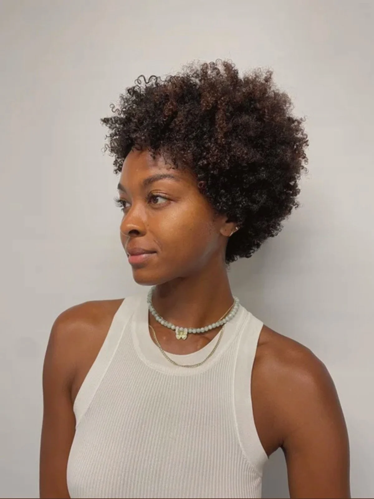 A woman with short curly dark hair, wearing a sleeveless white top, layered necklaces, and small earrings, standing against a plain light gray wall.