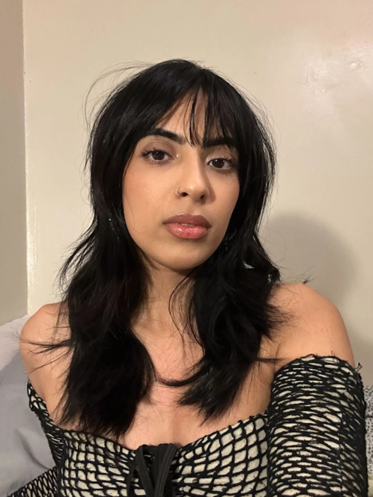 A woman with long black hair and a nose ring, wearing a black mesh top, is taking a selfie indoors against a plain, light-colored wall.