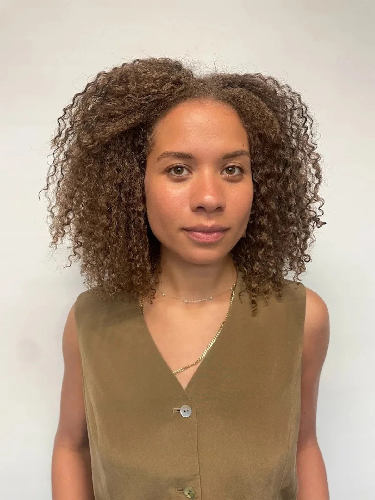 Portrait of a woman with curly hair wearing a sleeveless olive-green top and layered necklaces, standing against a plain white background.