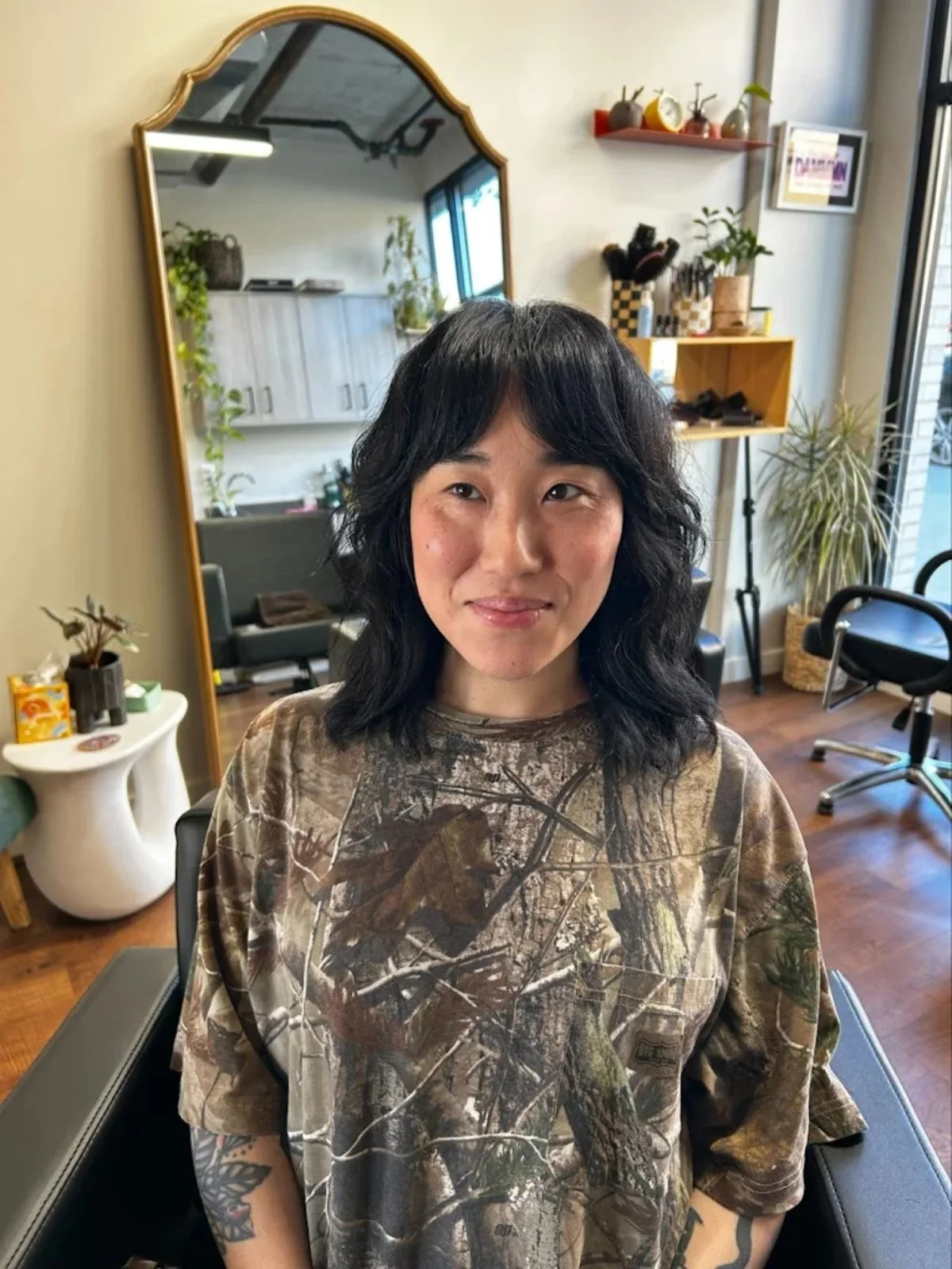 A woman with black wavy hair and tattoos on her right arm, smiling slightly, sitting in a salon chair with a background of a mirror, plants, and salon equipment.