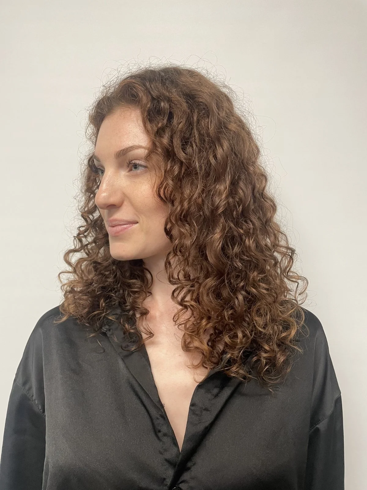 A woman with shoulder-length curly red hair and fair skin is wearing a black satin shirt and looking to her right against a plain white background.