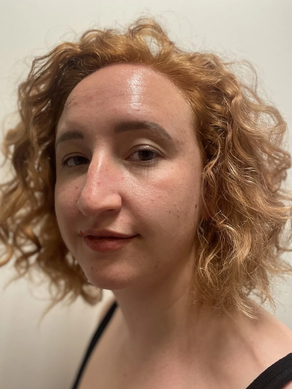 Close-up of woman with curly red hair, fair skin, light makeup, and a subtle smile, wearing a black top.
