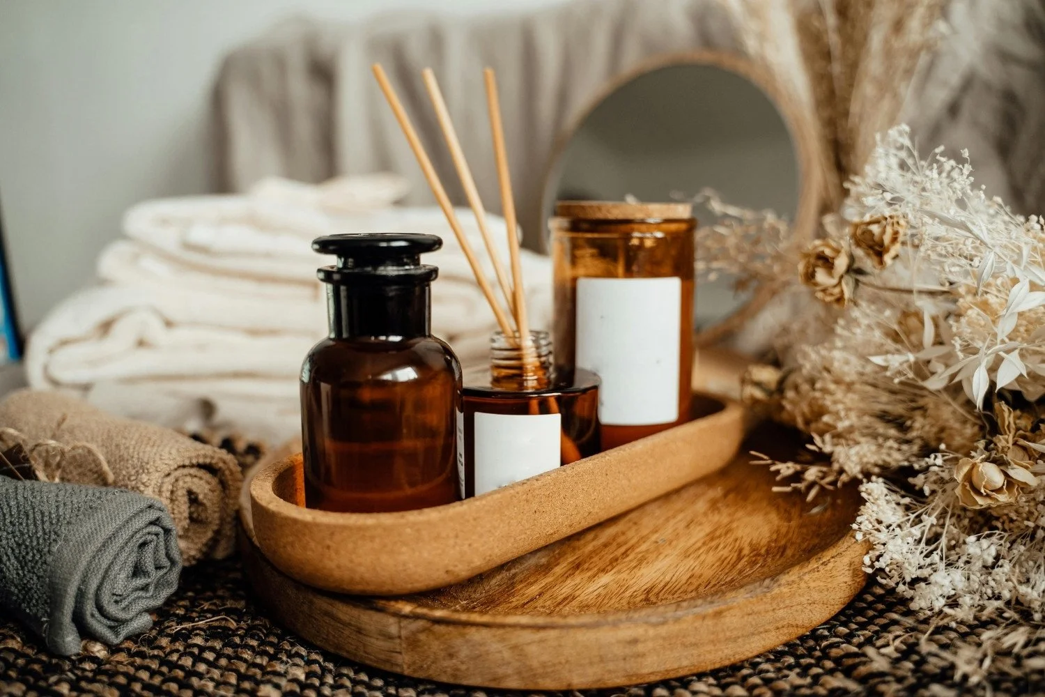 A wooden tray holding amber glass bottles, one with reed sticks, and a mirror. Dried flowers and rolled towels are around the tray.