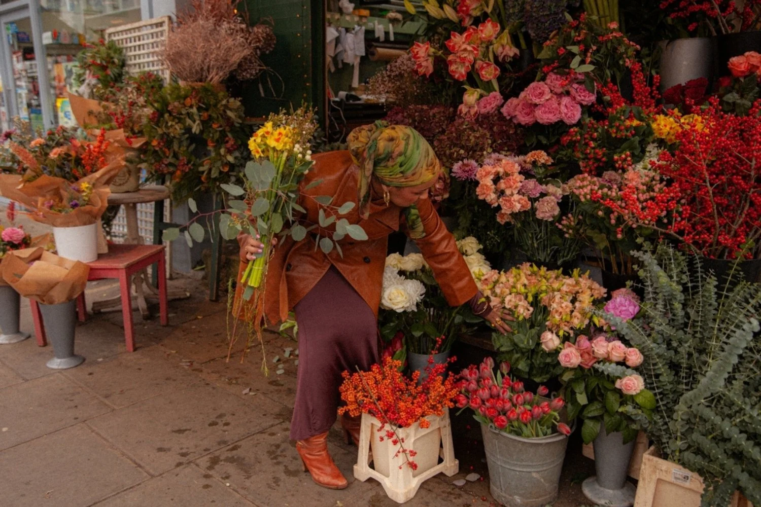 A woman selecting flowers at a flower shop on a city sidewalk. She is holding a bouquet of yellow flowers and is surrounded by various colorful flowers in pots and buckets.