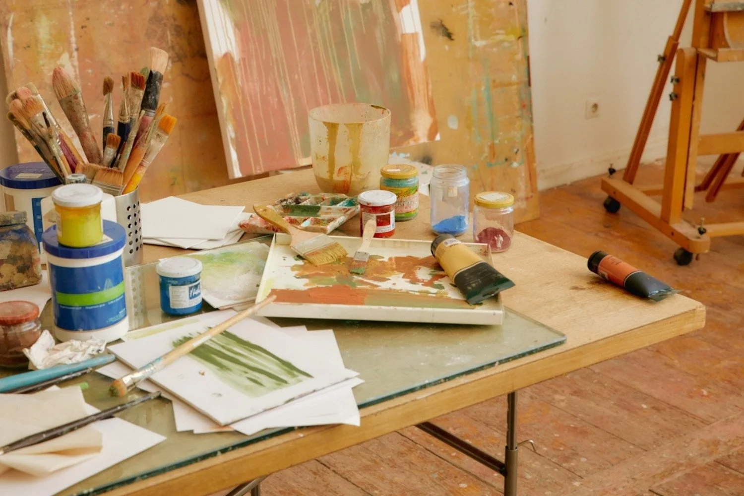 An artist's workspace with paint bottles, brushes, paper, and paintings on the table and walls.