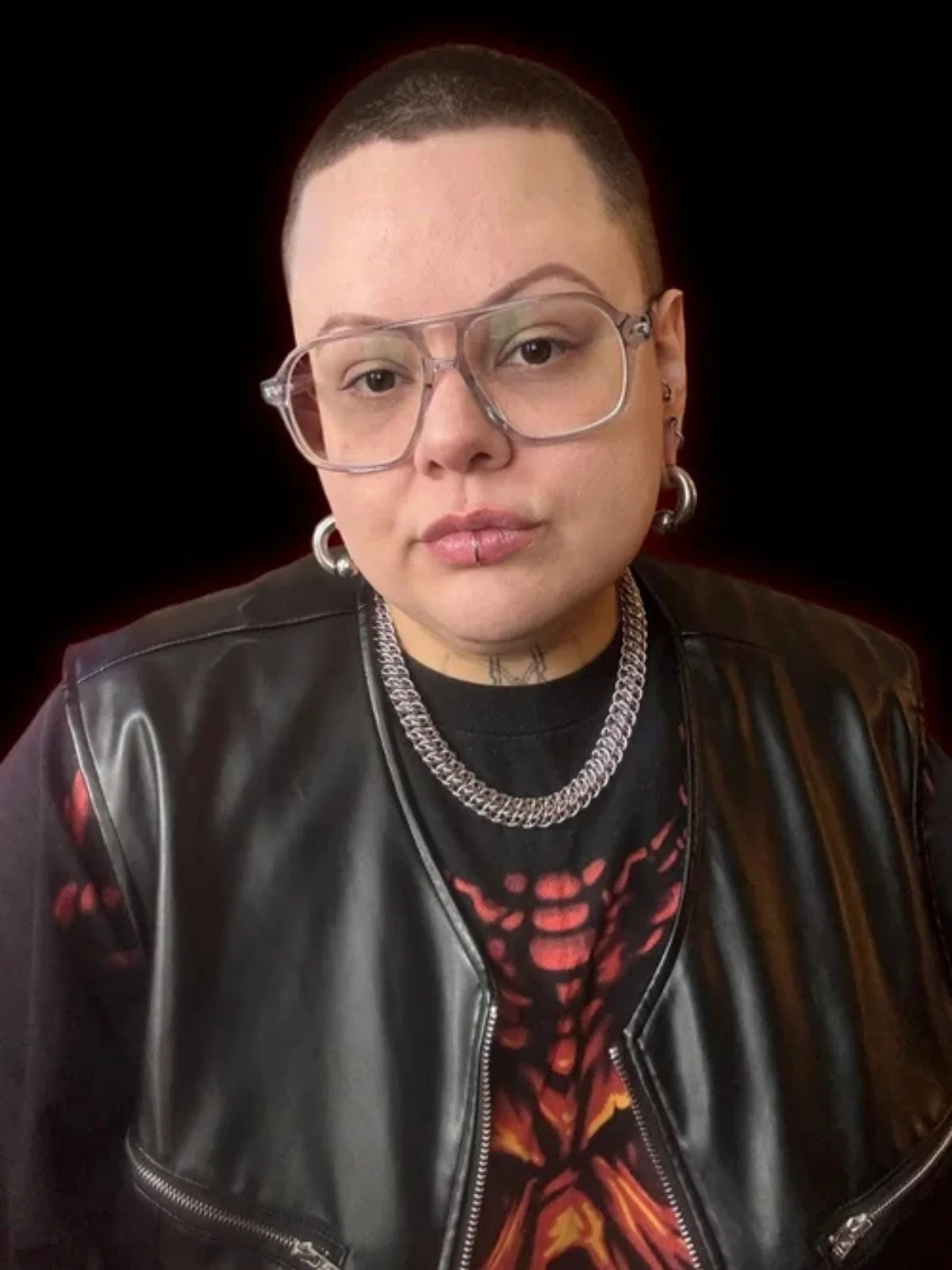 Person with short hair, wearing glasses, a leather vest, and silver jewelry, against a black background.