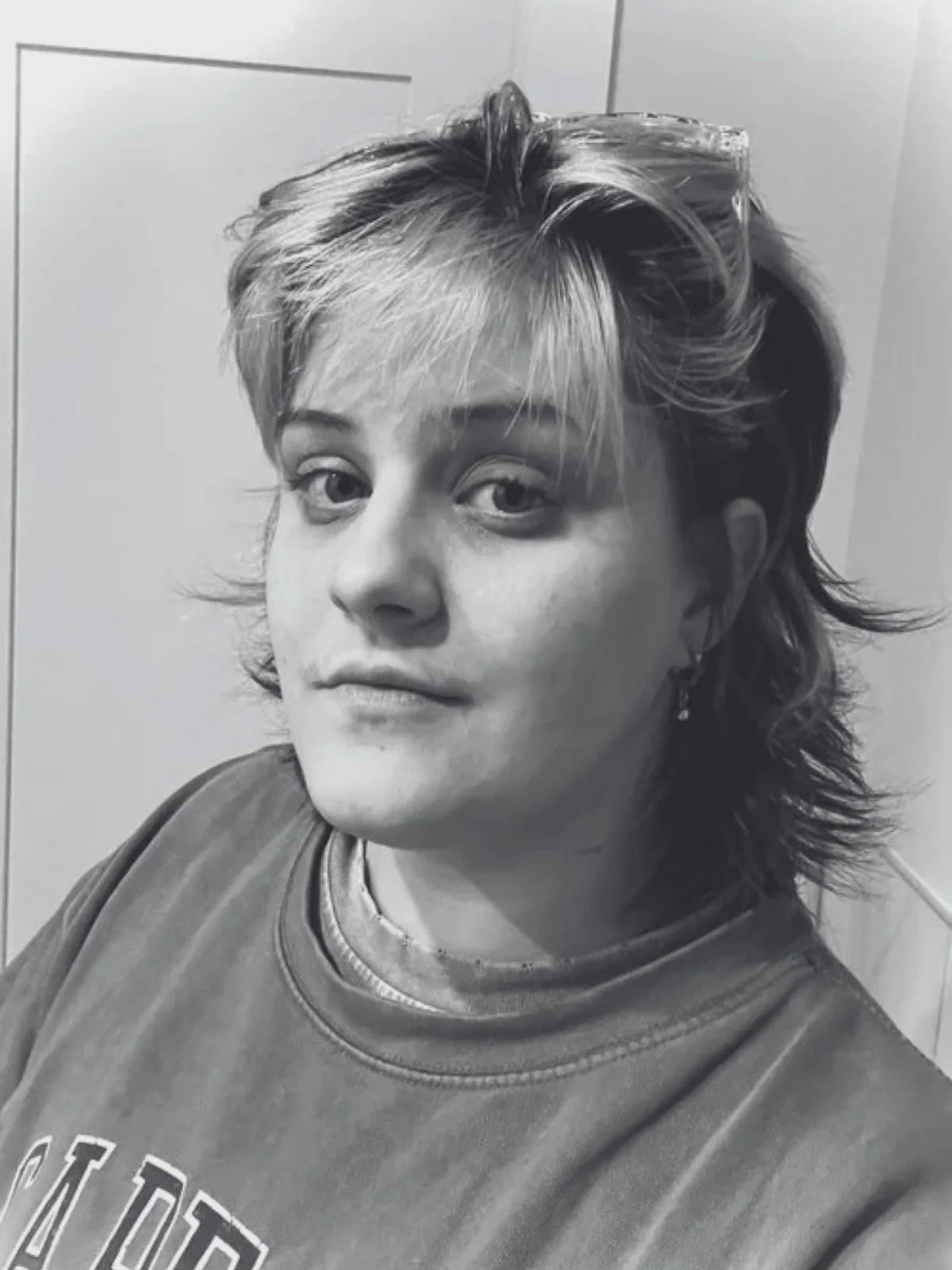 Black and white photo of a woman with short, layered, tousled hair, wearing a casual sweatshirt and earrings, looking at the camera.