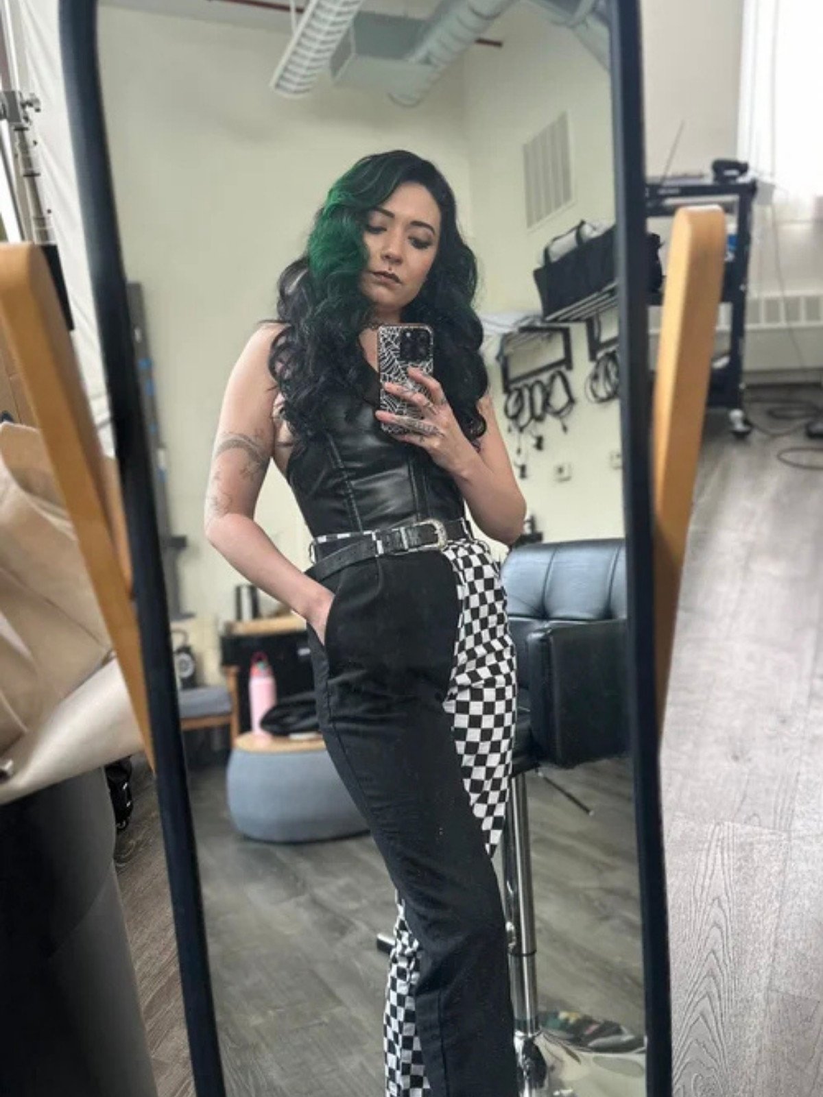 A woman taking a mirror selfie in a room, with dark wavy hair that has green highlights, wearing a black sleeveless top and checkered black and white pants.