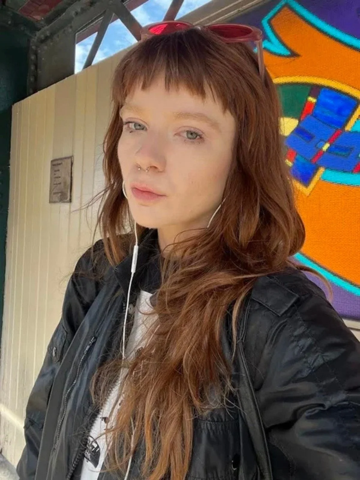 A young woman with long, wavy red hair, wearing a black jacket and red sunglasses on her head, taking a selfie at an outdoor location with a colorful sign in the background.