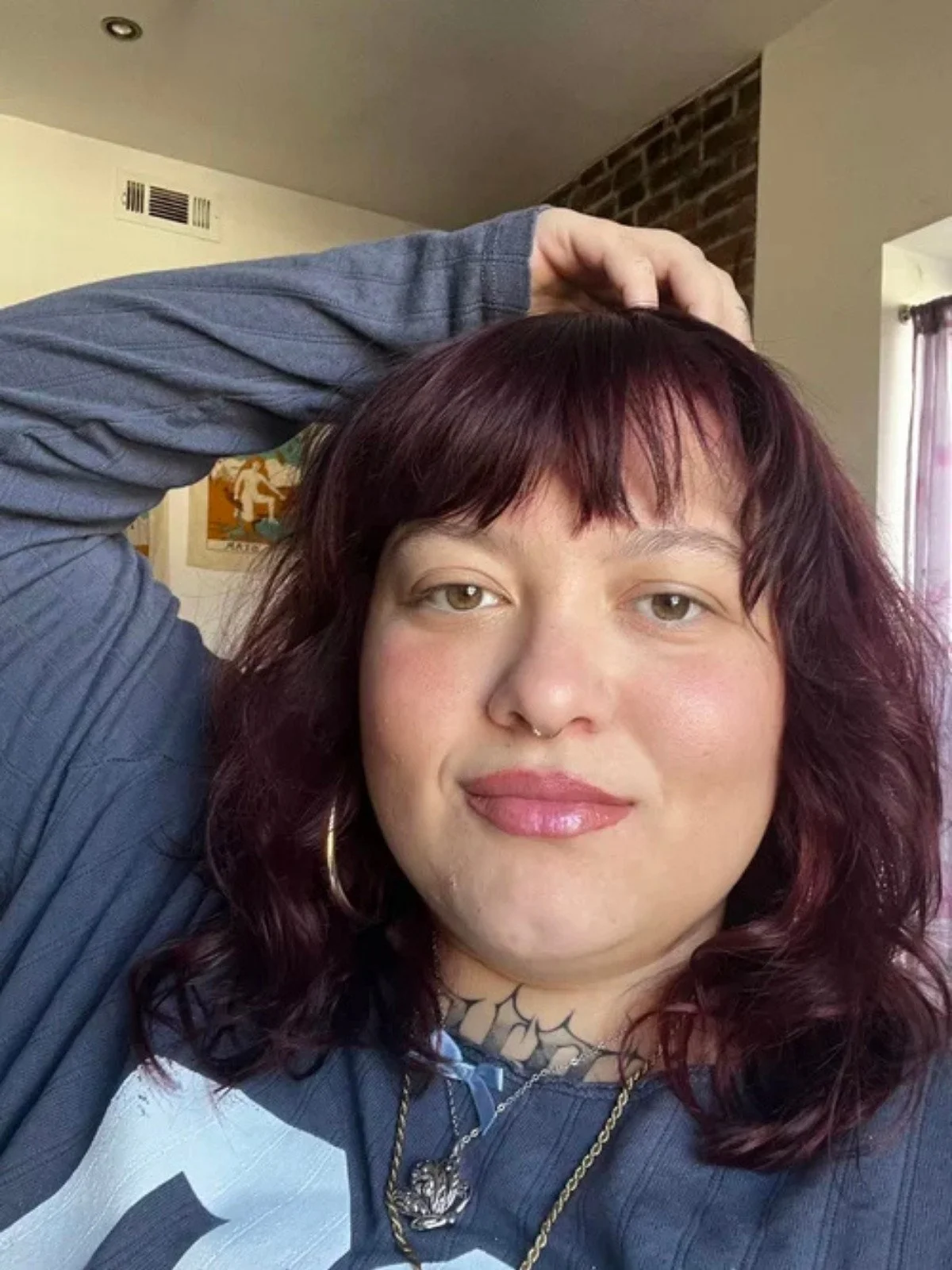 A woman with shoulder-length wavy burgundy hair, light skin, and brown eyes, smiling and wearing hoop earrings, a nose ring, a necklace, and a blue and white shirt, inside a room with a brick wall and a window with curtains.