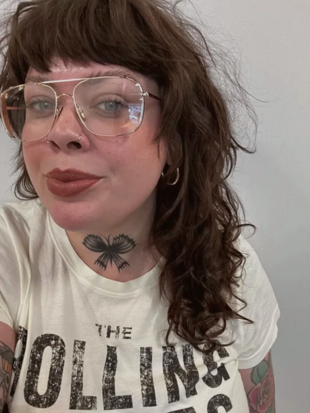 A woman with curly brown hair, glasses, and tattoos on her neck and arms. She is wearing a white T-shirt and hoop earrings, and is standing against a plain gray wall.
