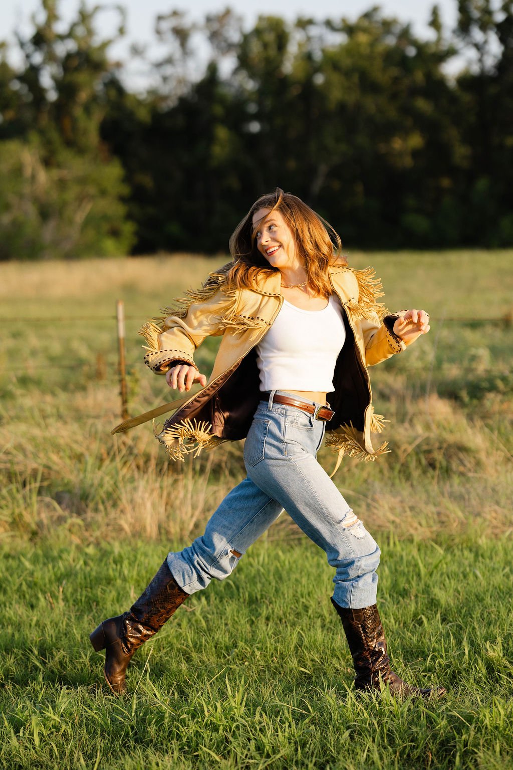A woman running in a grassy field during the daytime, wearing a fringed tan jacket, white crop top, ripped jeans, and cowboy boots, smiling and looking to the side.