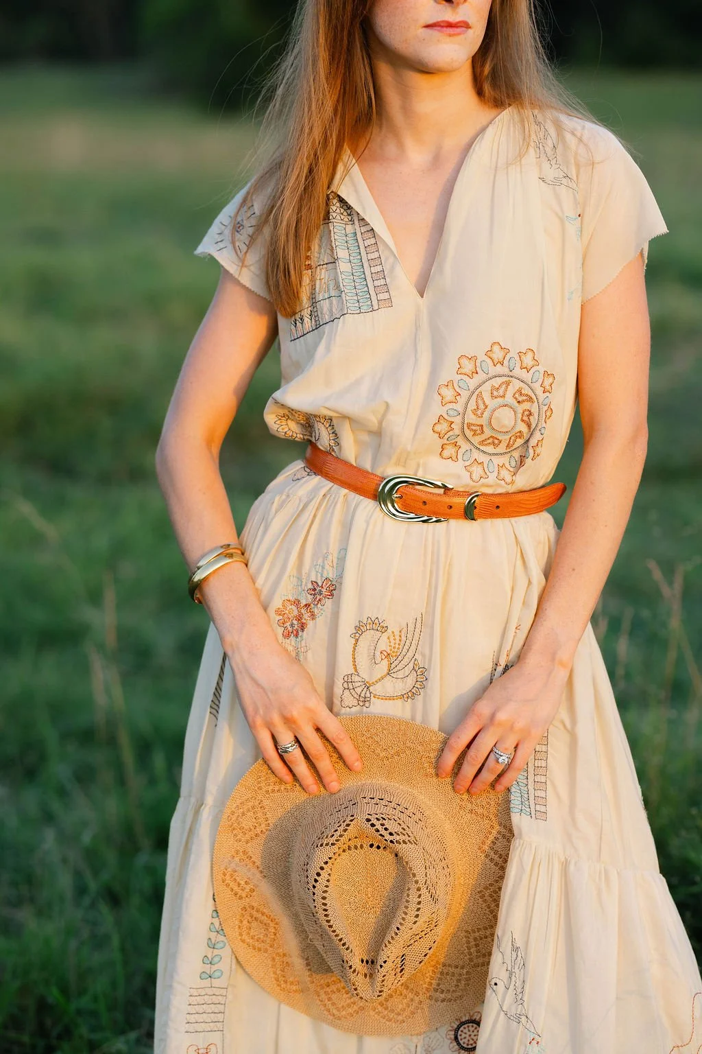 Woman in a light-colored embroidered dress holding a straw hat in a grassy outdoor setting.