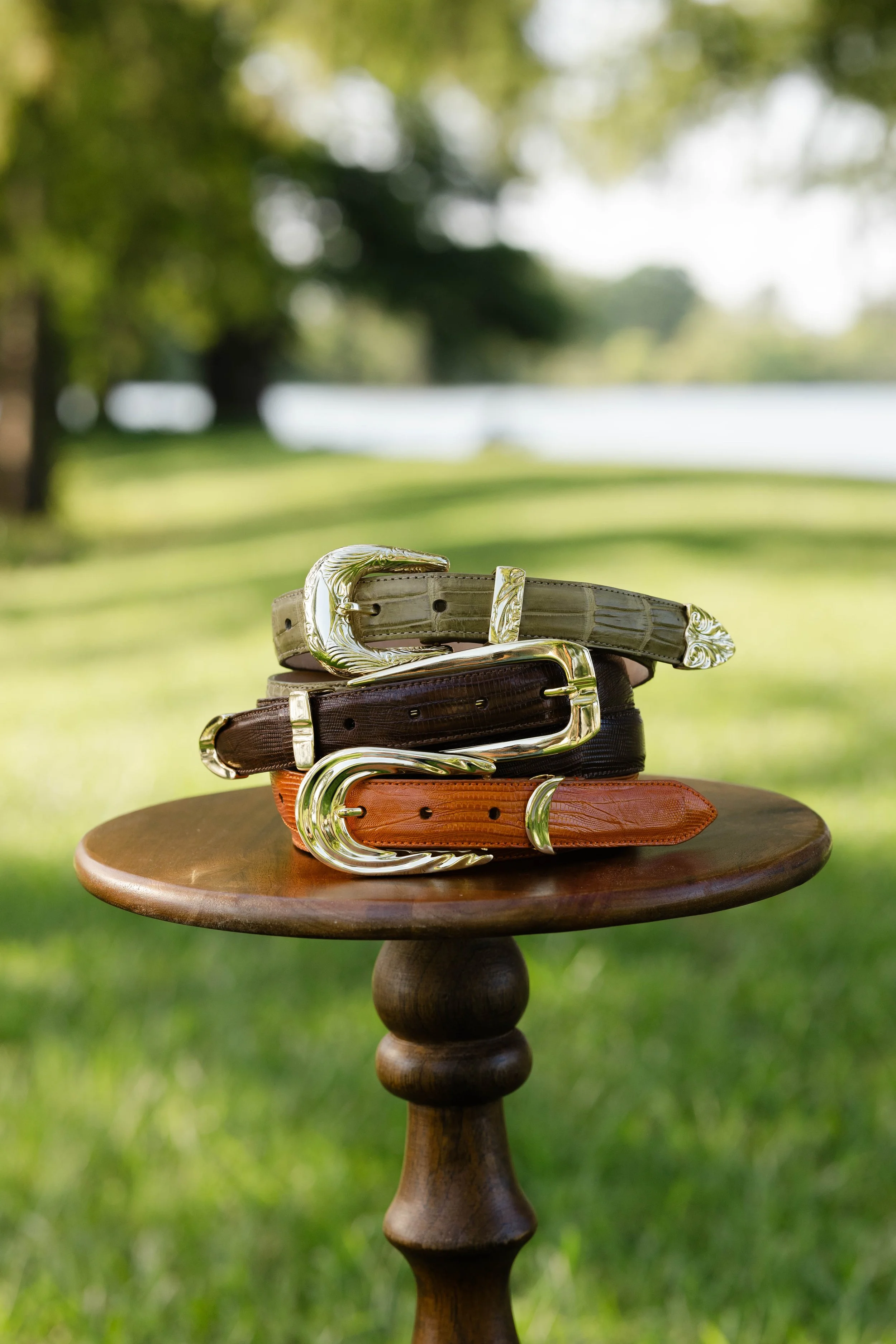 Stacked leather belts with gold buckles on a wooden table outdoors, blurred green trees and water in the background.